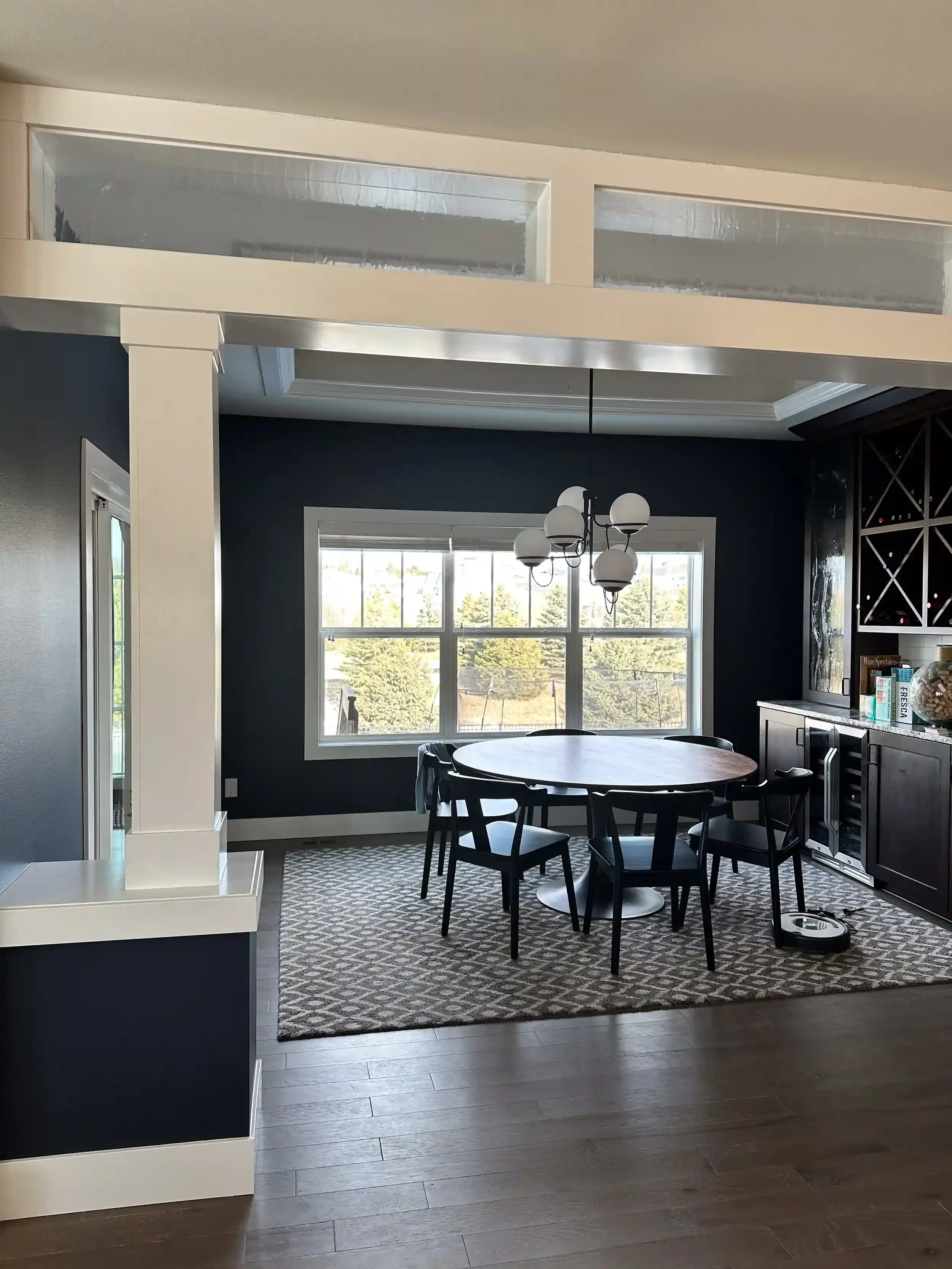 View of a dining area with a round black table, four black chairs, a window with a view of trees, a chandelier with five spherical lights, a dark blue accent wall, a patterned area rug, and a kitchen side with a built-in wine rack and counters.