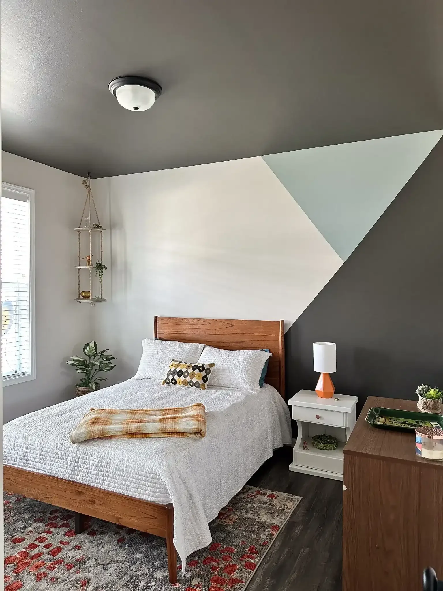 A cozy bedroom with a wooden bed, white quilt, and decorative pillows, a white nightstand with a lamp, a potted plant, and a shelf with plants and ornaments. The wall behind the bed is painted in white and dark gray with a geometric pattern, and a window with blinds is visible on the left side.