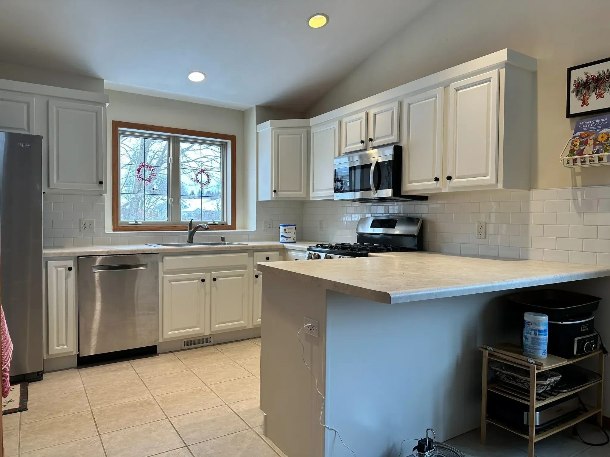 A kitchen with white cabinets, a window with a view of trees outside, and a beige countertop. There is a stainless steel microwave above the stove, a dishwasher, and various small appliances and decorations.