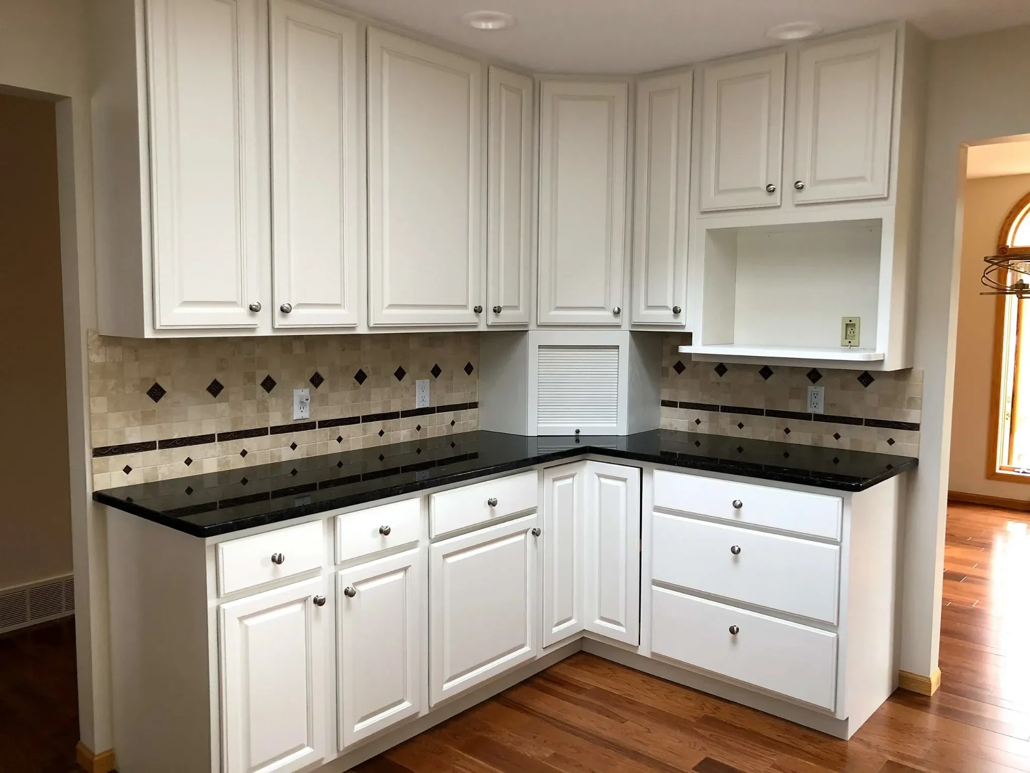 White kitchen cabinets with silver knobs, beige tiled backsplash with small diamond accents, black granite countertops, and hardwood floors.