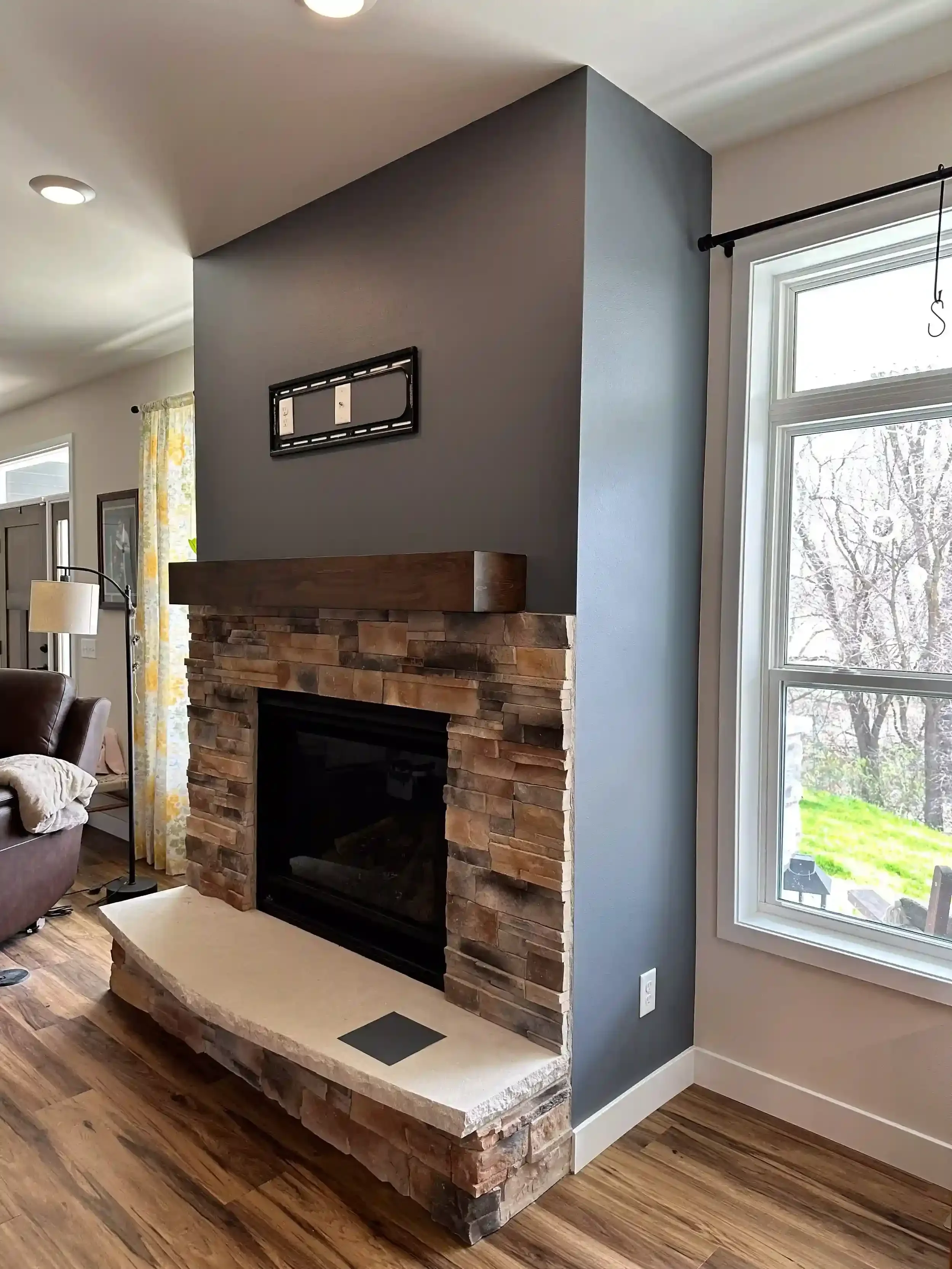 Living room with a brick fireplace, large window with a view of trees outside, and a dark gray wall. The fireplace has a beige stone hearth and a wood mantle. Part of a brown armchair and lamp are visible on the left.