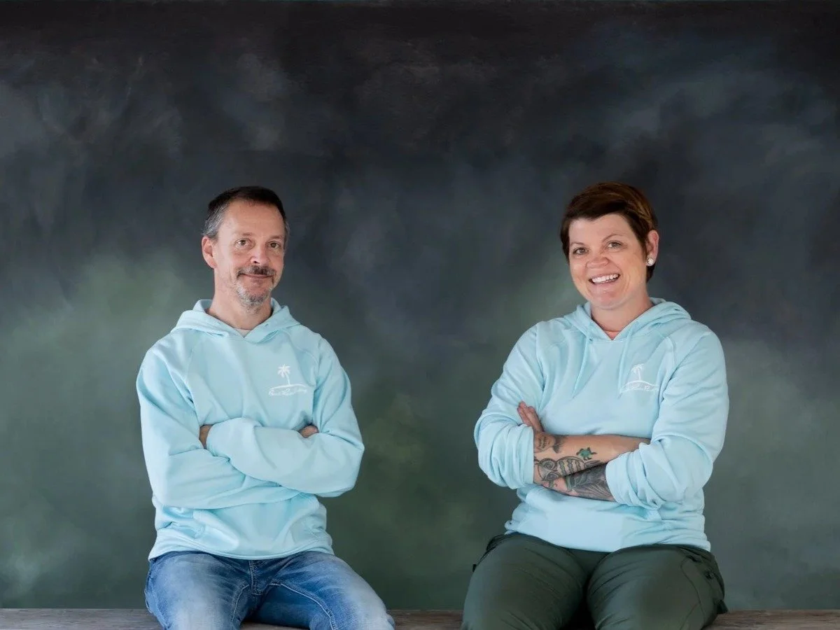 A man and a woman sitting on a wooden surface in front of a cloudy sky background, both wearing light blue hoodies with a white logo, smiling at the camera.