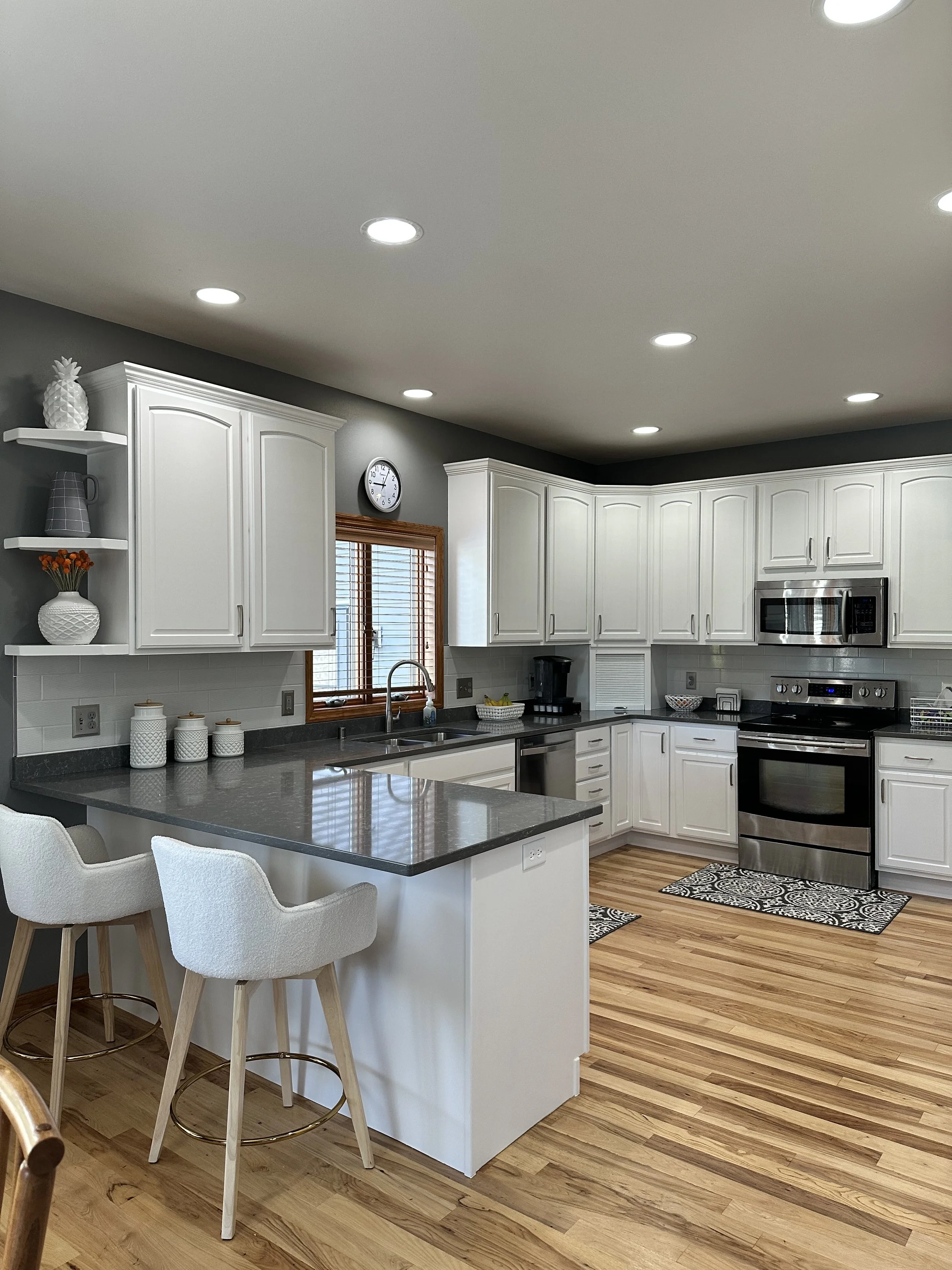 Modern kitchen with white cabinetry, stainless steel appliances, dark countertop, light hardwood flooring, gray walls, and two white upholstered chairs at a kitchen island.