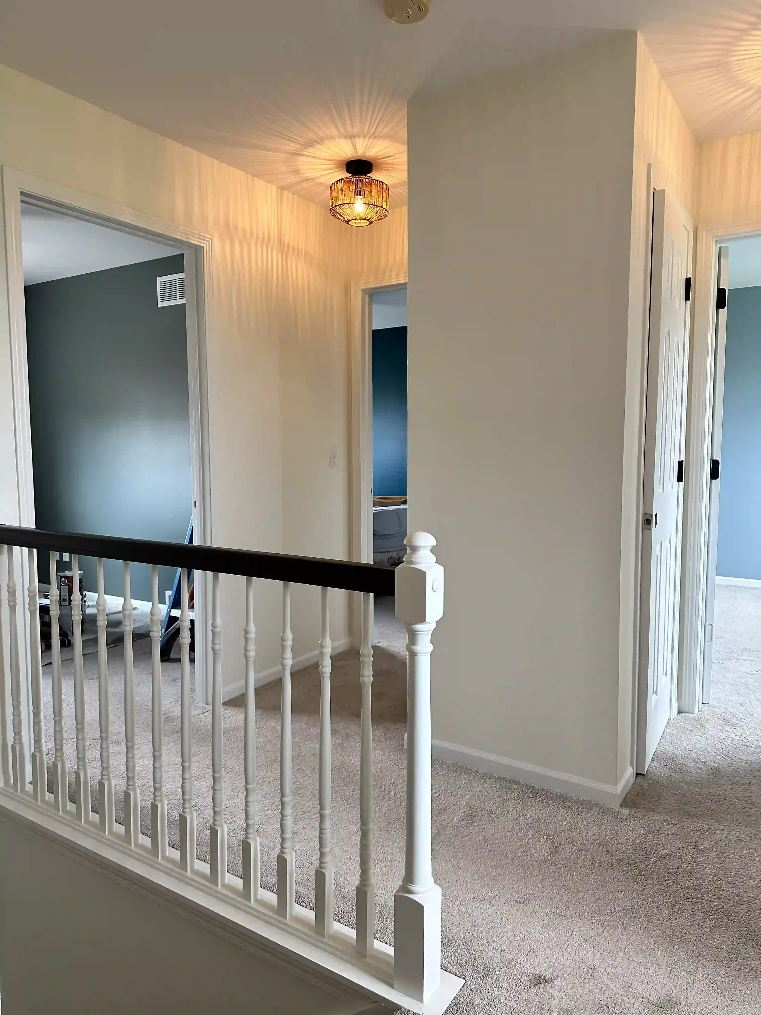 Interior view of a house with a hallway, multiple doorways, a staircase with a white railing, beige carpeted floors, a wall-mounted light fixture, and light-colored walls.