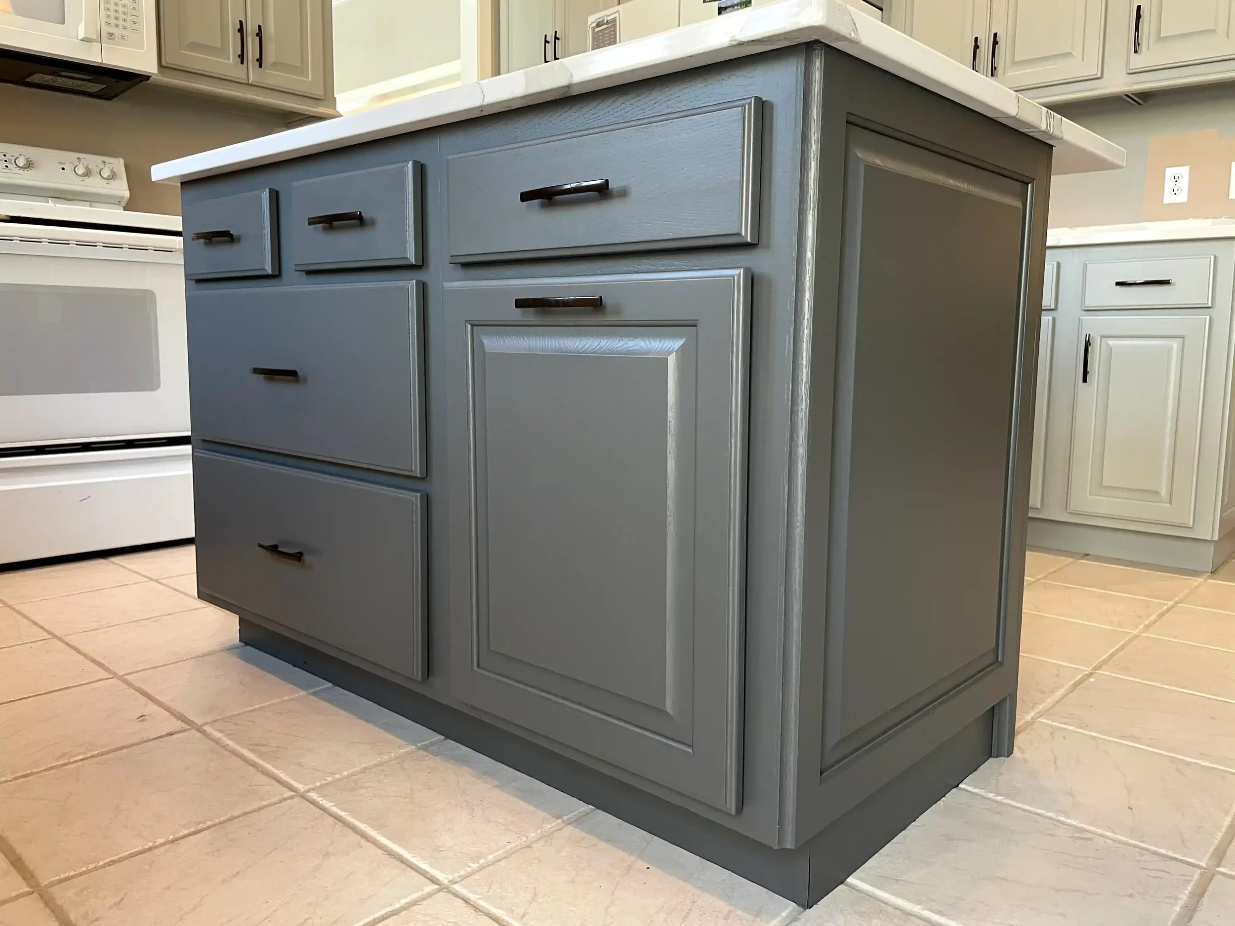 A kitchen island painted in light gray with multiple drawers and cabinet doors, topped with a white countertop, situated in a kitchen with white cabinets and a stove in the background.