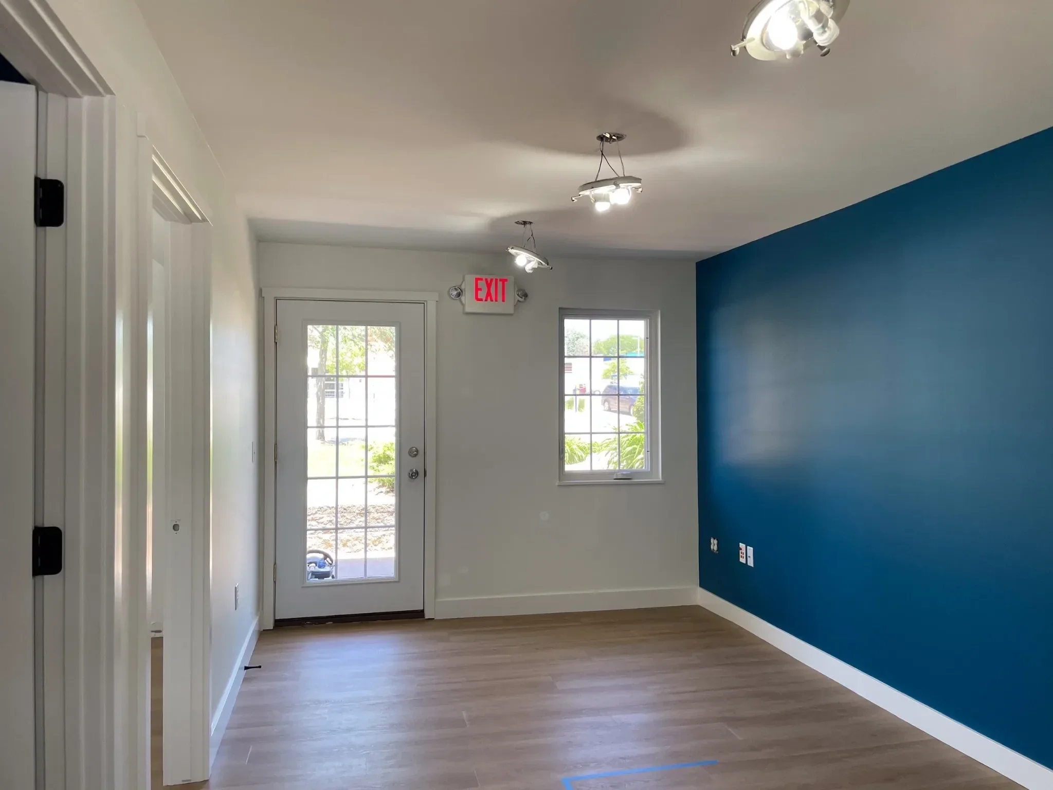 Interior of a room with white walls, a blue accent wall, light hardwood floors, a door with glass panels, windows letting in natural light, ceiling lights, and a visible electrical outlet.