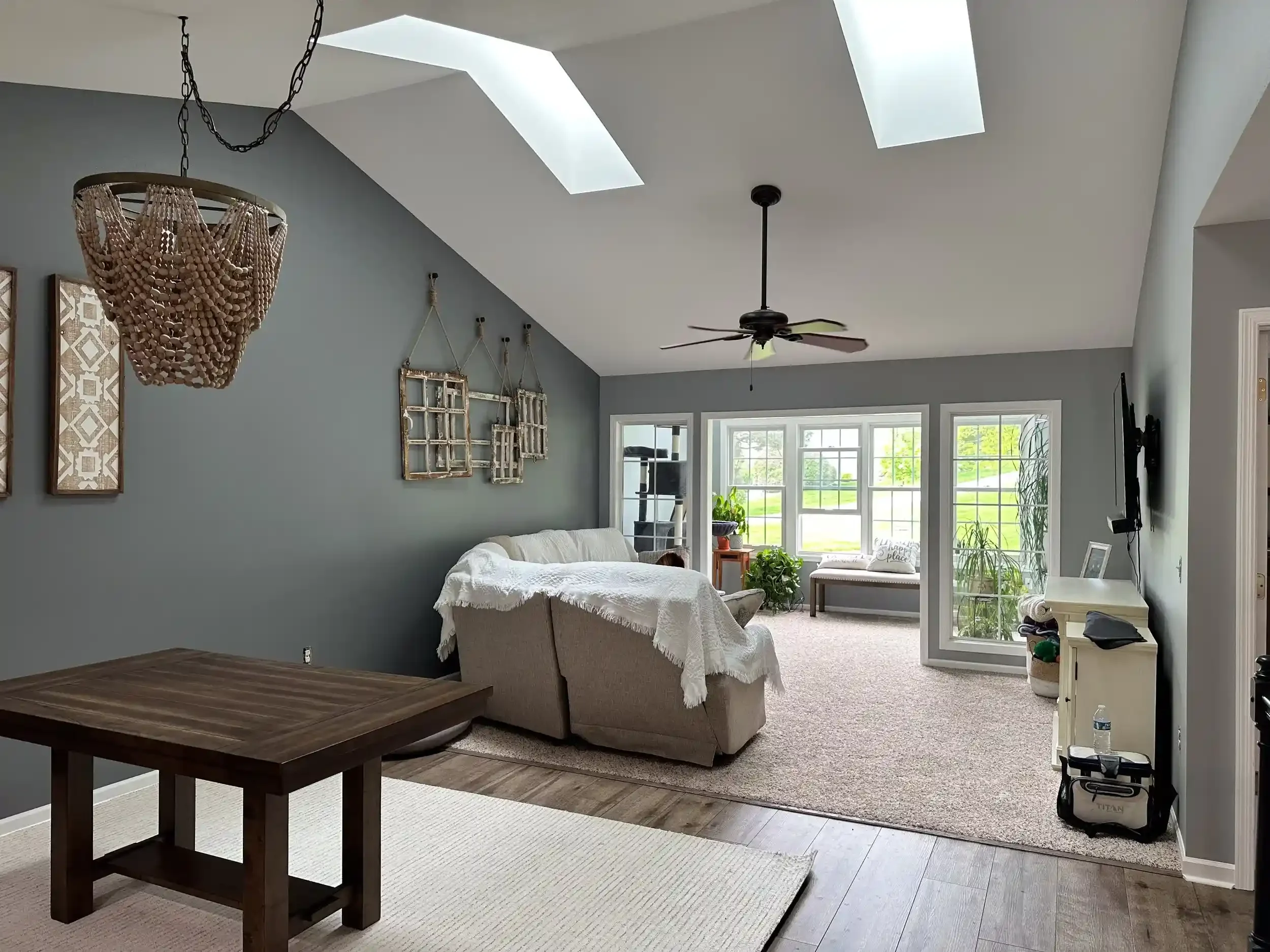 Open concept living room with gray walls, skylights, ceiling fan, beige sofa with white cover, decorative wall mirrors, plants, and a large window facing outside.