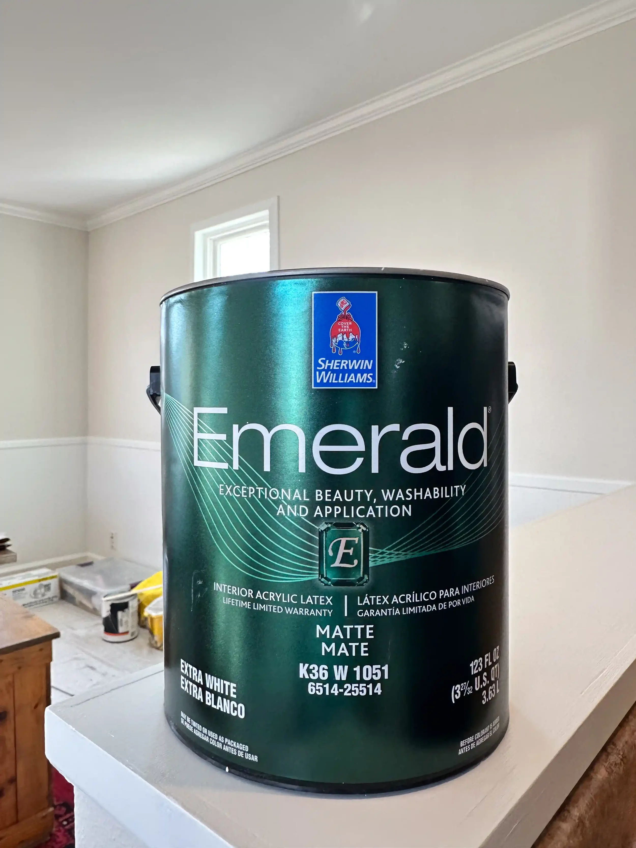 Close-up of a large can of Sherwin Williams Emerald exterior paint labeled in English and Spanish, placed on a white surface in a room under renovation.
