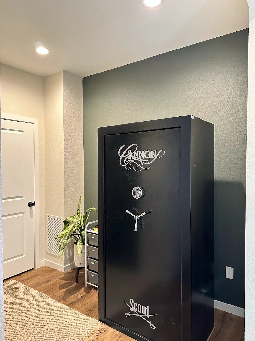 A large black safe with the words "Cannon Scout" painted on it, located in a room with beige walls, a white door, and a small plant in a white pot.