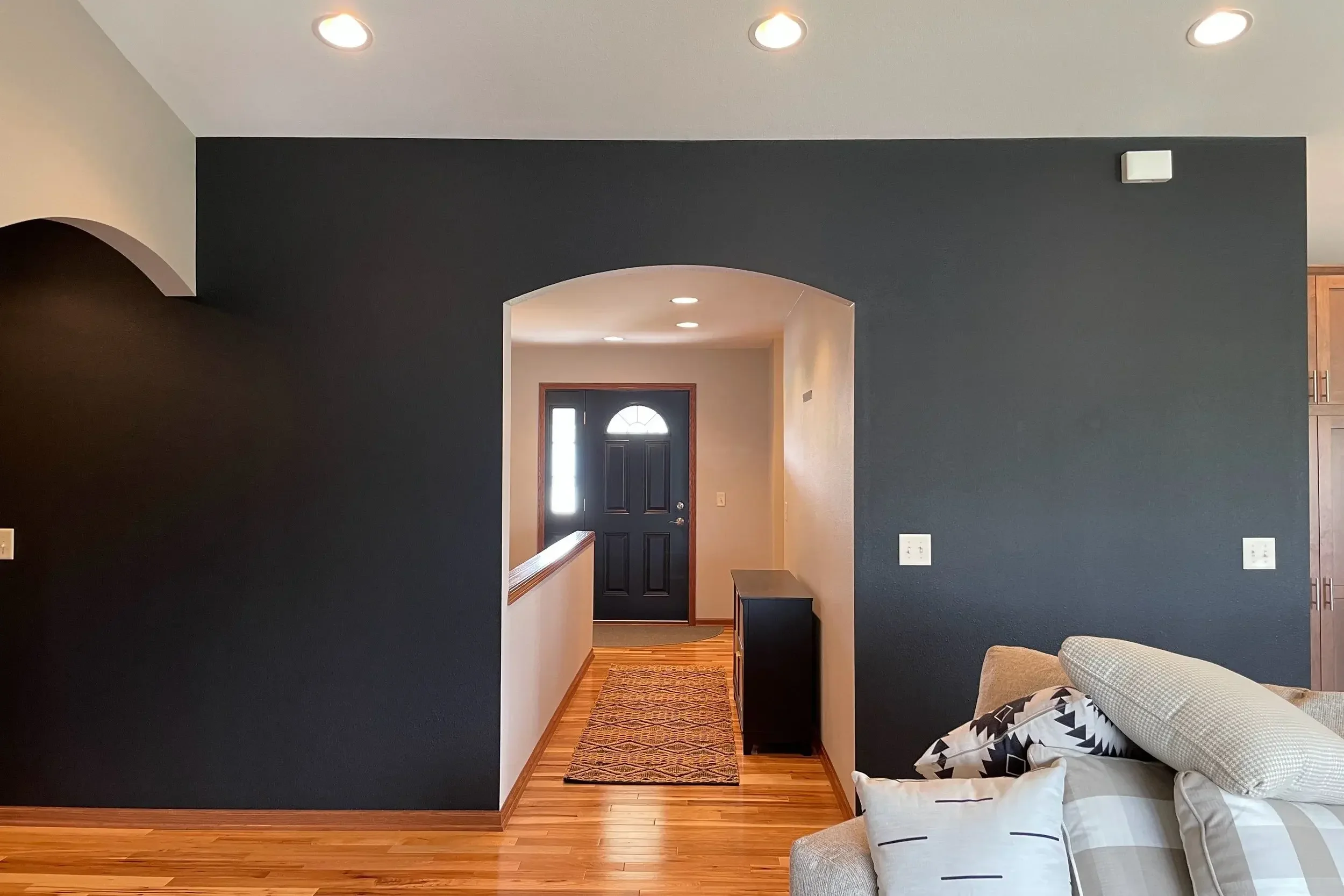 Interior view of a home entrance with a black front door, visible through an archway, with a hallway, hardwood floors, and a patterned rug. The living room has a dark accent wall, a beige sectional sofa with patterned pillows, and nearby wooden cabin
