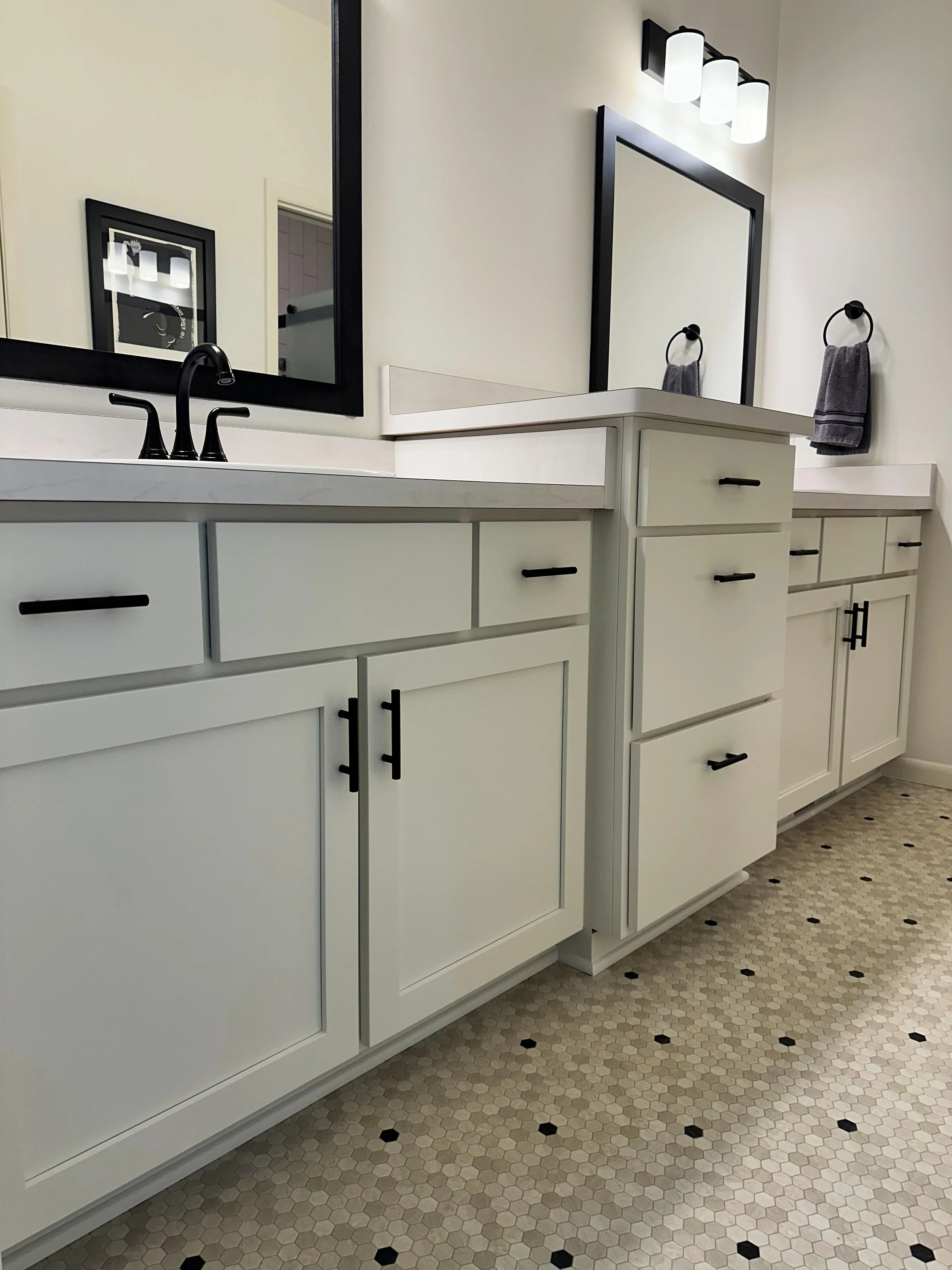 A bathroom vanity with white cabinets, black handles, and a black faucet. Two mirrors are visible, one large standard and a smaller framed one. There is a black towel ring with a gray towel, and a modern light fixture with four white lamps above the 