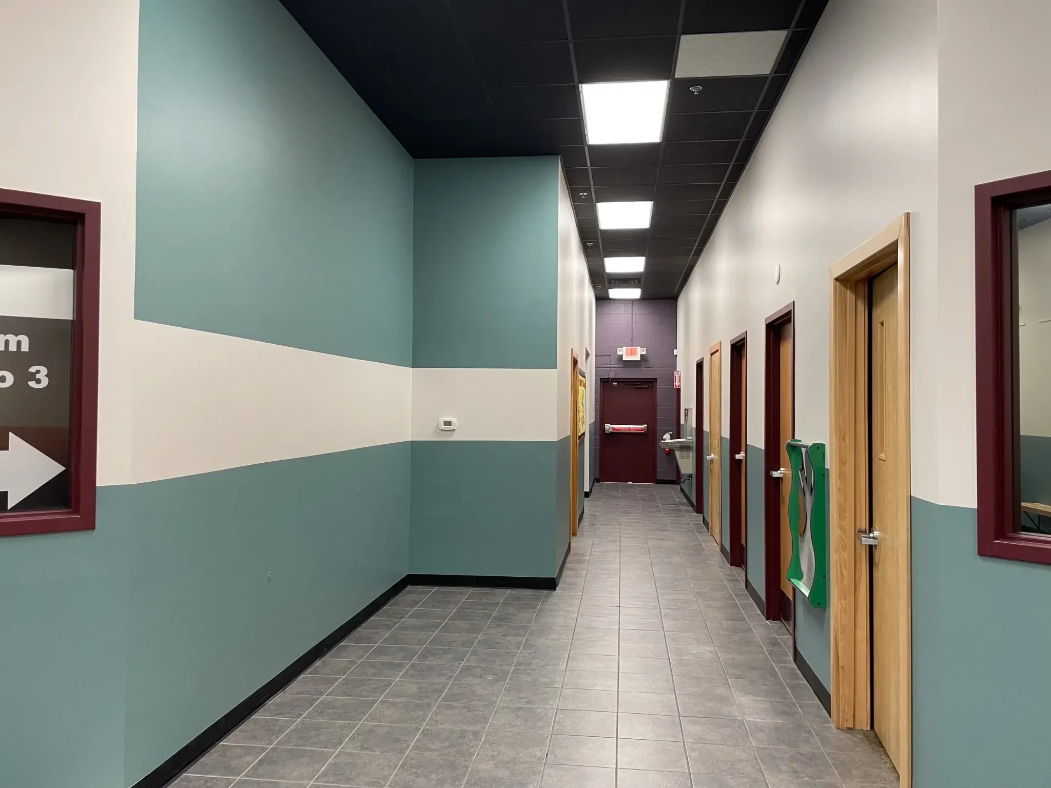 A hallway with a teal and white painted wall, tile flooring, multiple wooden doors on the right, and fluorescent ceiling lights.