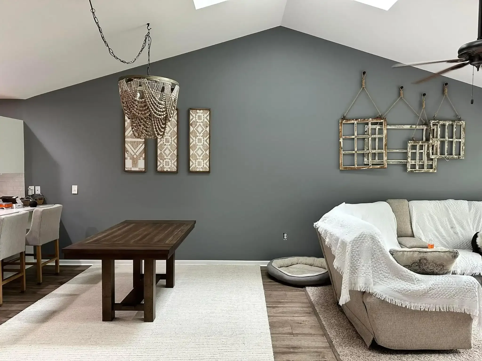 Living room with gray walls, hanging decorations, a ceiling fan, a couch with a white blanket, a dog bed, and a wooden dining table with chairs.
