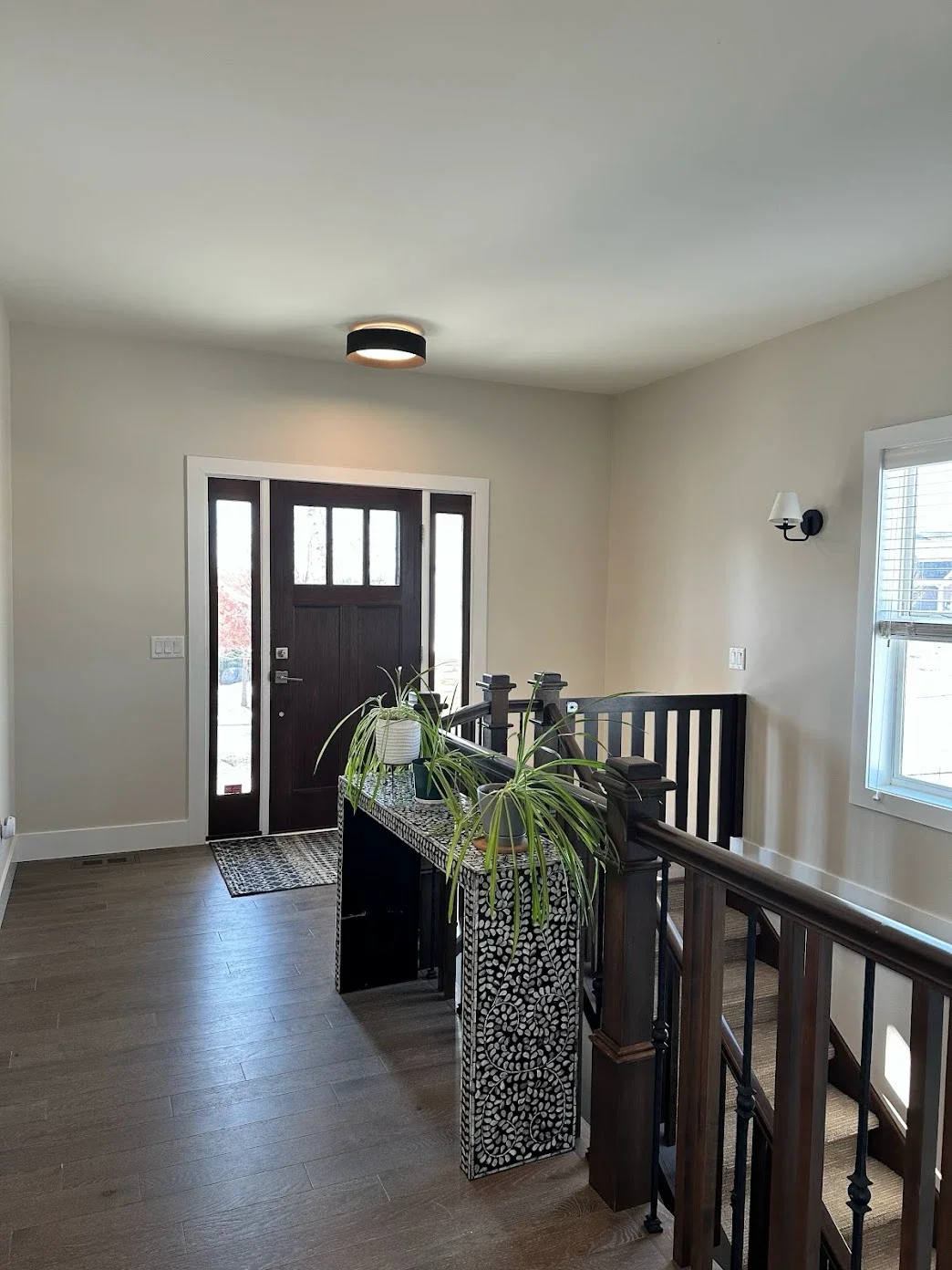 Entryway with front door, window, and staircase. Indoor plants on a decorative black and white stand. Modern ceiling and wall lights. Hardwood floors and neutral-colored walls.