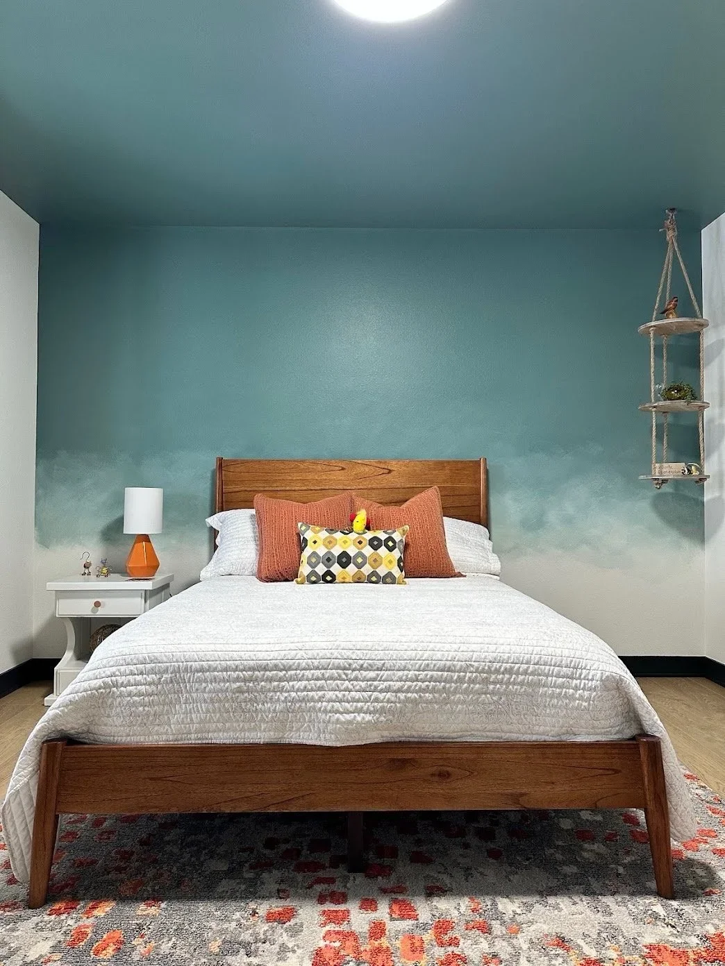 Cozy bedroom with a wooden bed, white quilt, orange and patterned pillows, nightstand with white and orange lamp, teal accent wall, and hanging shelf with decor.