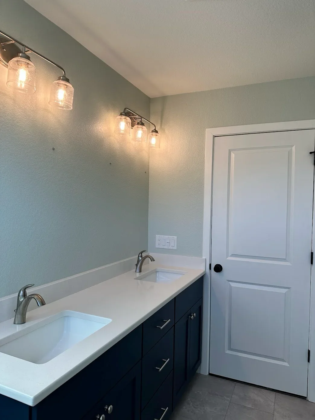 Bathroom with a dual-sink white countertop, dark blue cabinets, a white door, and modern glass-encased light fixtures.