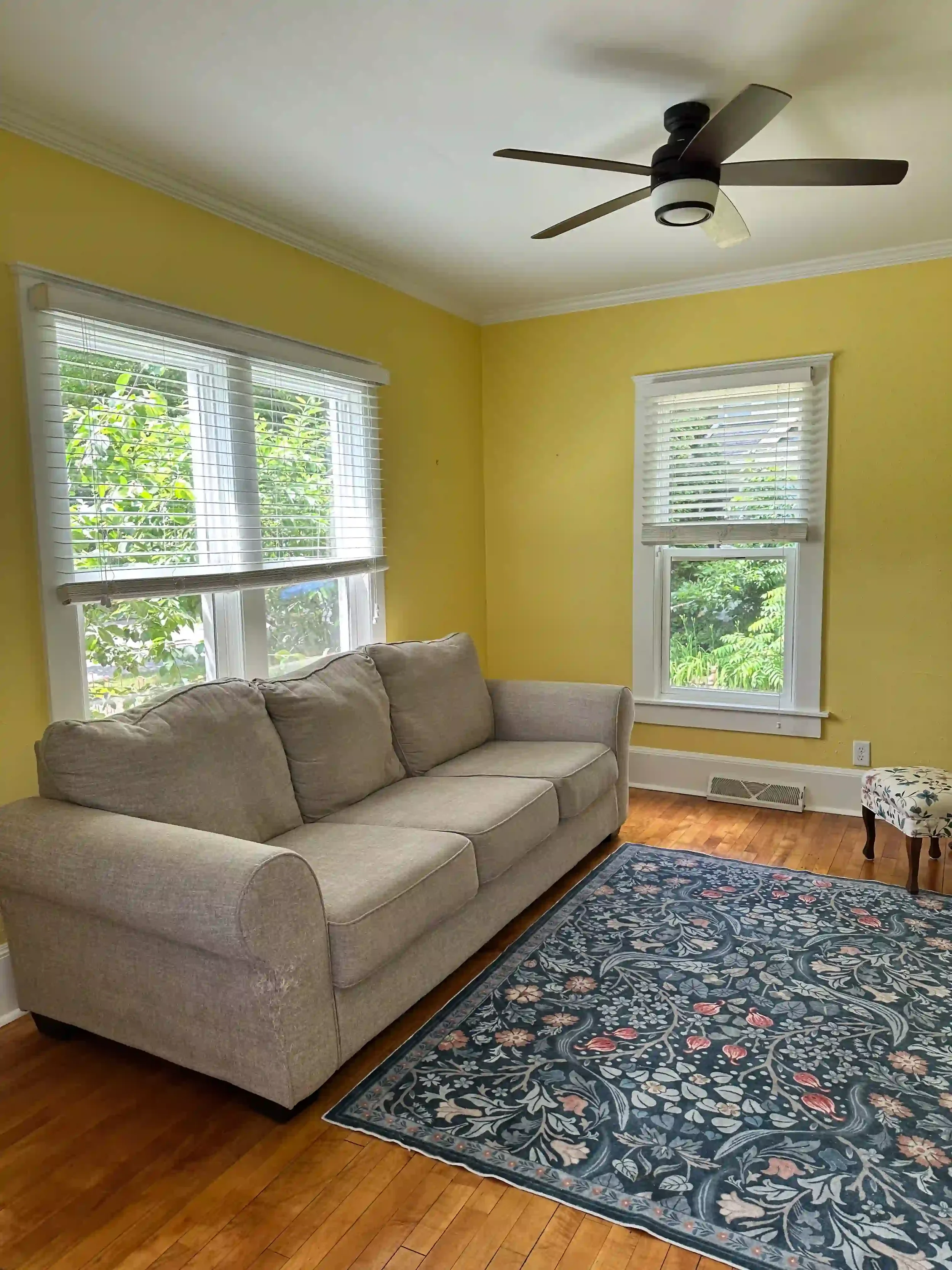 Living room with yellow walls, white trim, hardwood floor, beige sofa, floral rug, two windows with white blinds, ceiling fan with black blades, small floral upholstered stool.