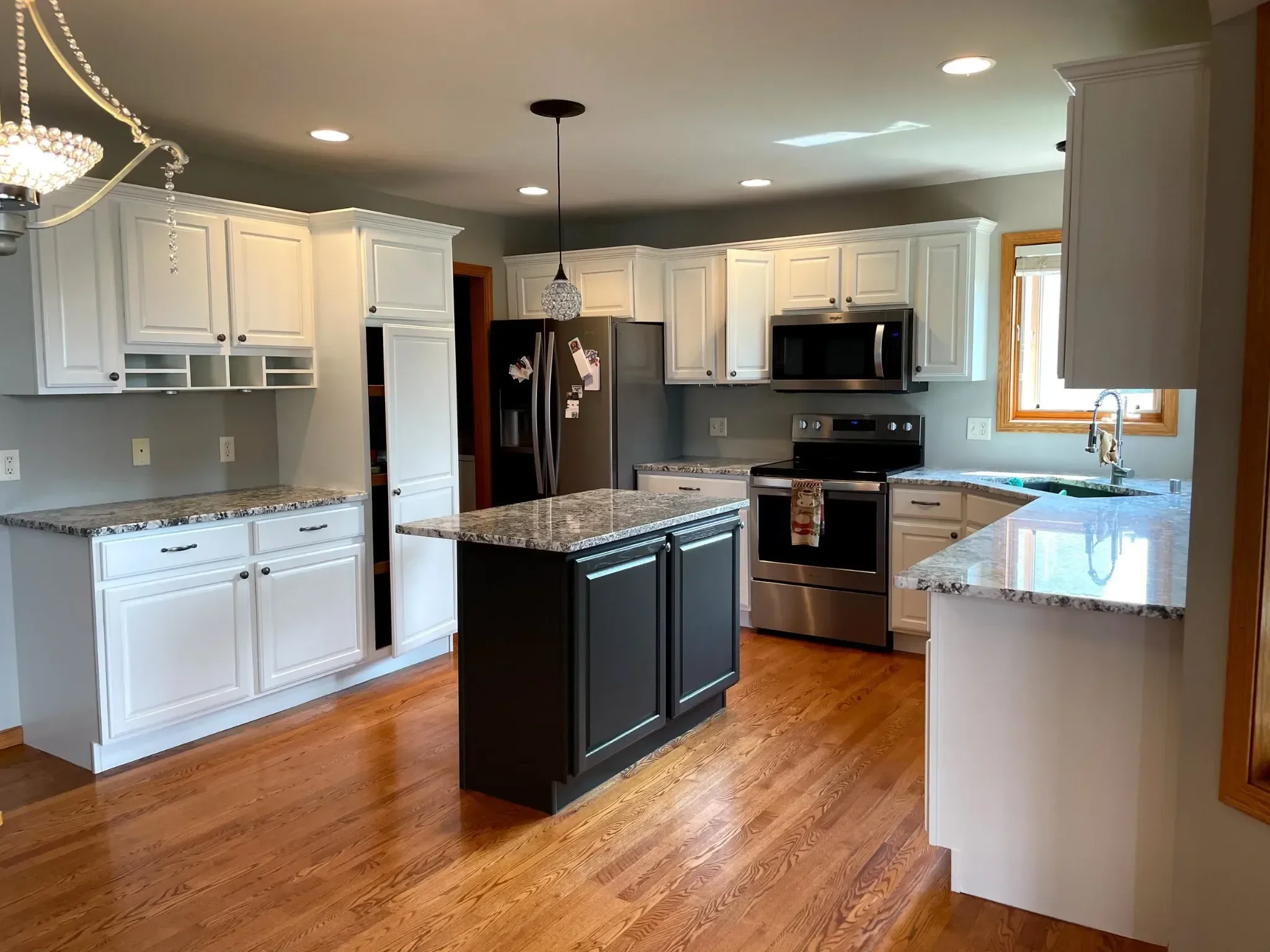 Modern kitchen with white cabinets, black island, granite countertops, hardwood floors, stainless steel appliances, and a window over the sink.