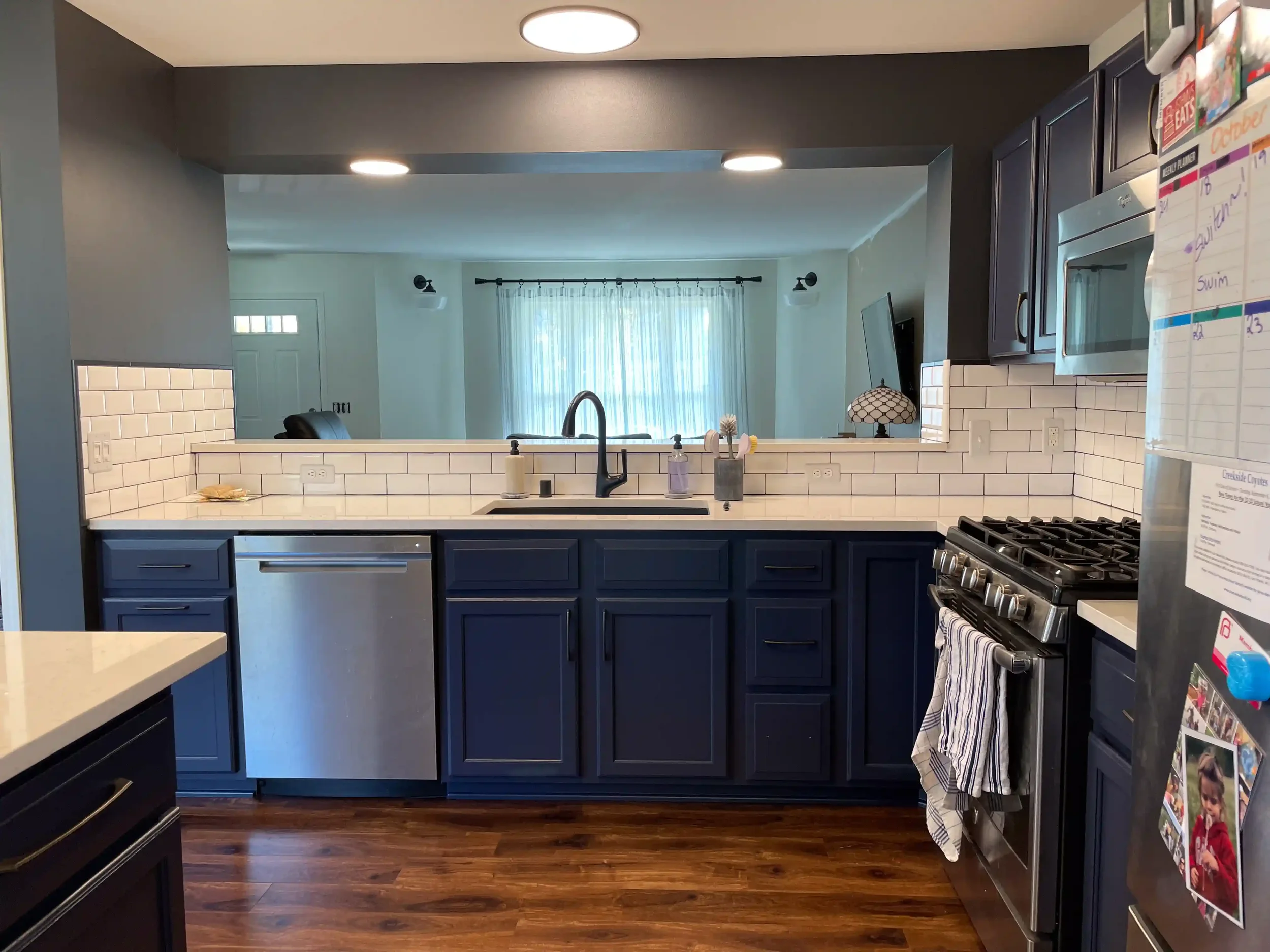 A kitchen with blue cabinets, white subway tile backsplash, and stainless steel appliances including a microwave and oven. There are dark wood floors and a view into a living room with a window and sheer curtains.