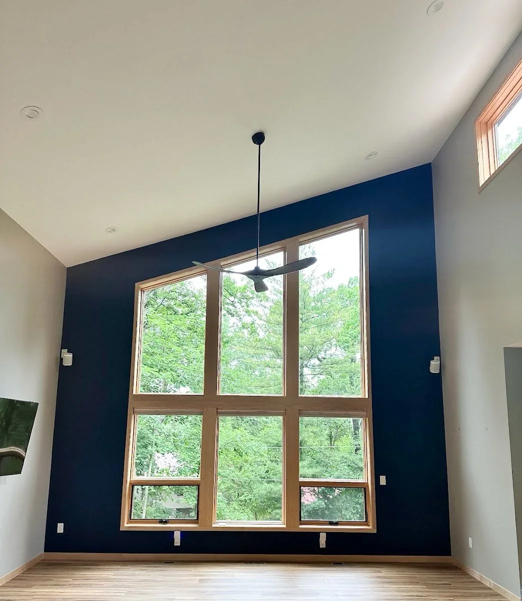 Empty living room with large wooden-framed window wall, ceiling fan, and hardwood floors, with green trees visible outside.