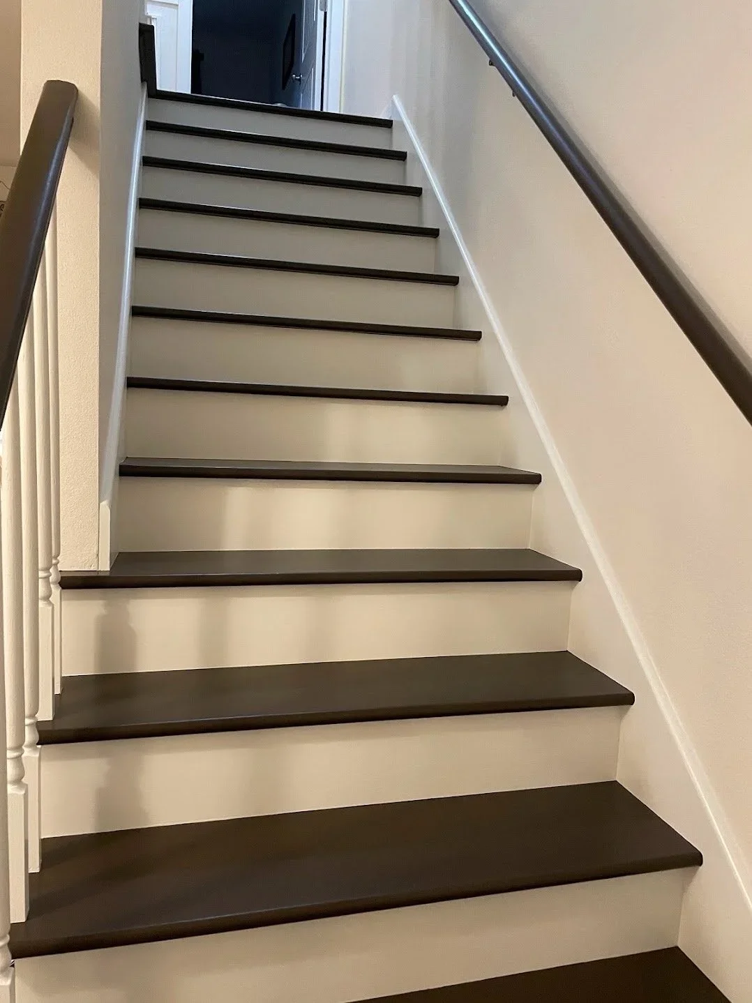 An indoor staircase with dark brown treads and white risers, with a dark brown handrail on the right side, leading up to an open doorway.