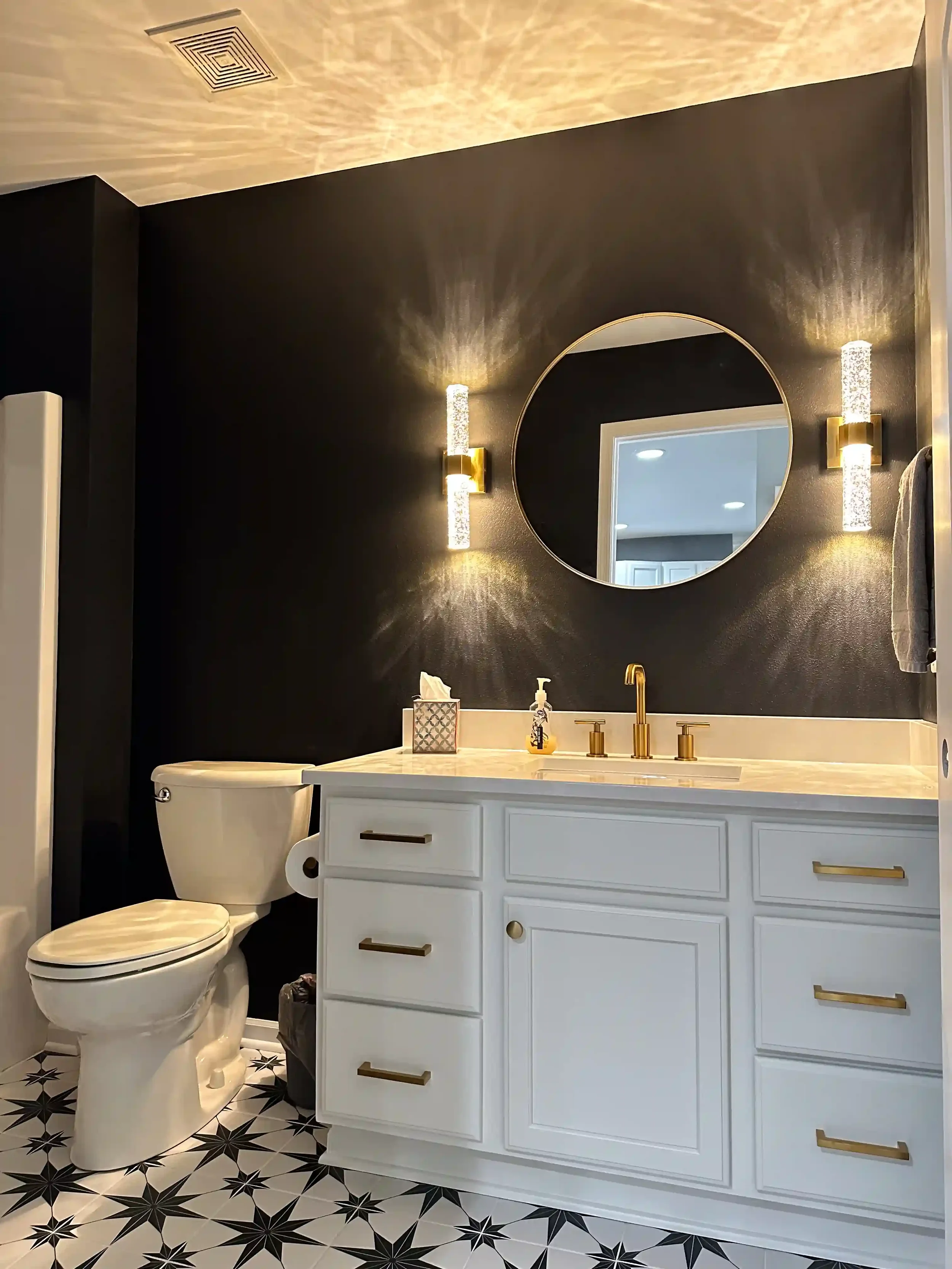 Modern bathroom with black and white star-patterned floor, white vanity with gold handles, round mirror, black wall, gold light fixtures, porcelain toilet, tissue box on vanity, soap dispenser, door visible in mirror reflection.