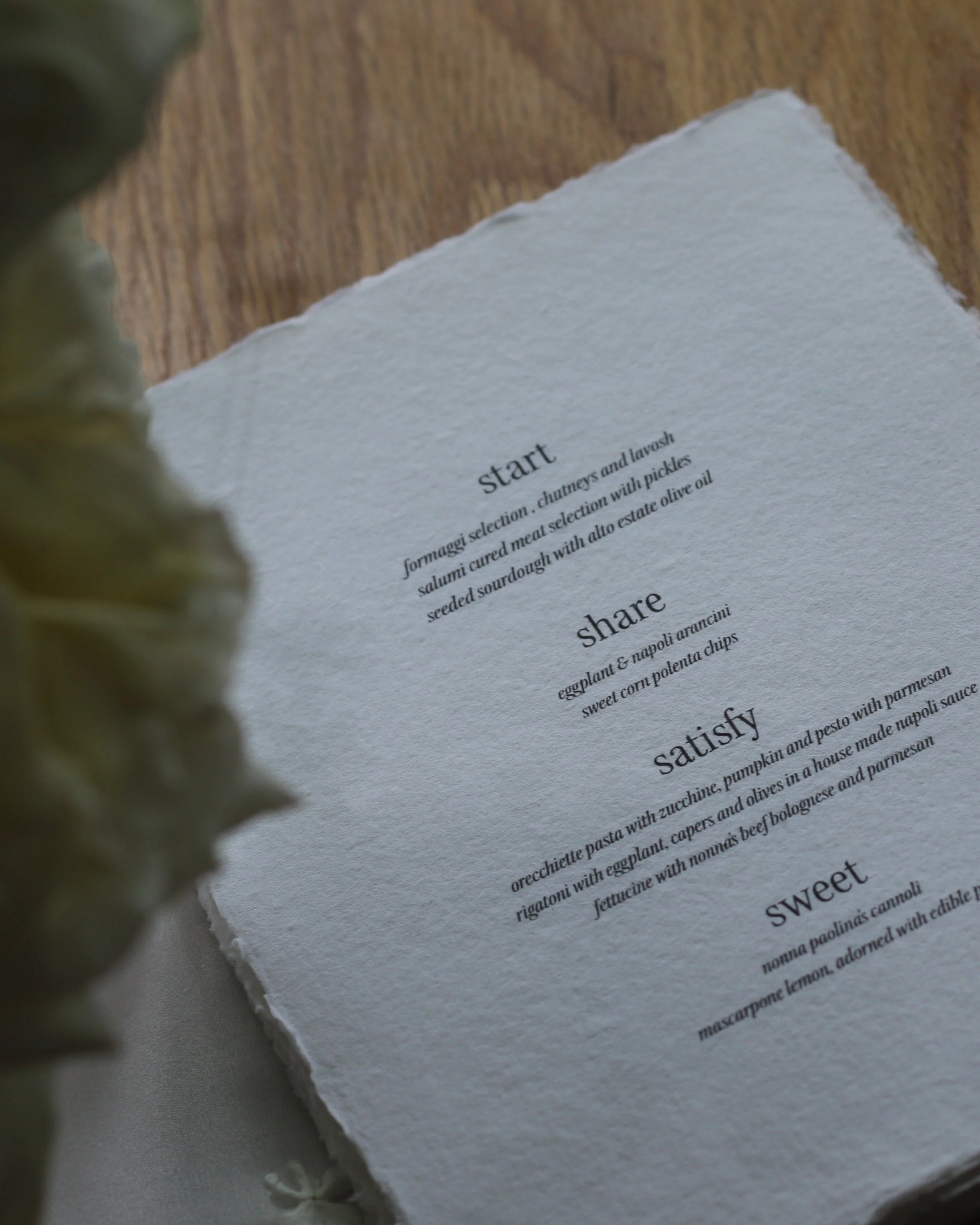 Close-up of a menu on textured paper featuring sections labeled 'start', 'share', 'satisfy', and 'sweet' with descriptions of dishes including cheese and meat selections, arancini, pasta, and cannoli.