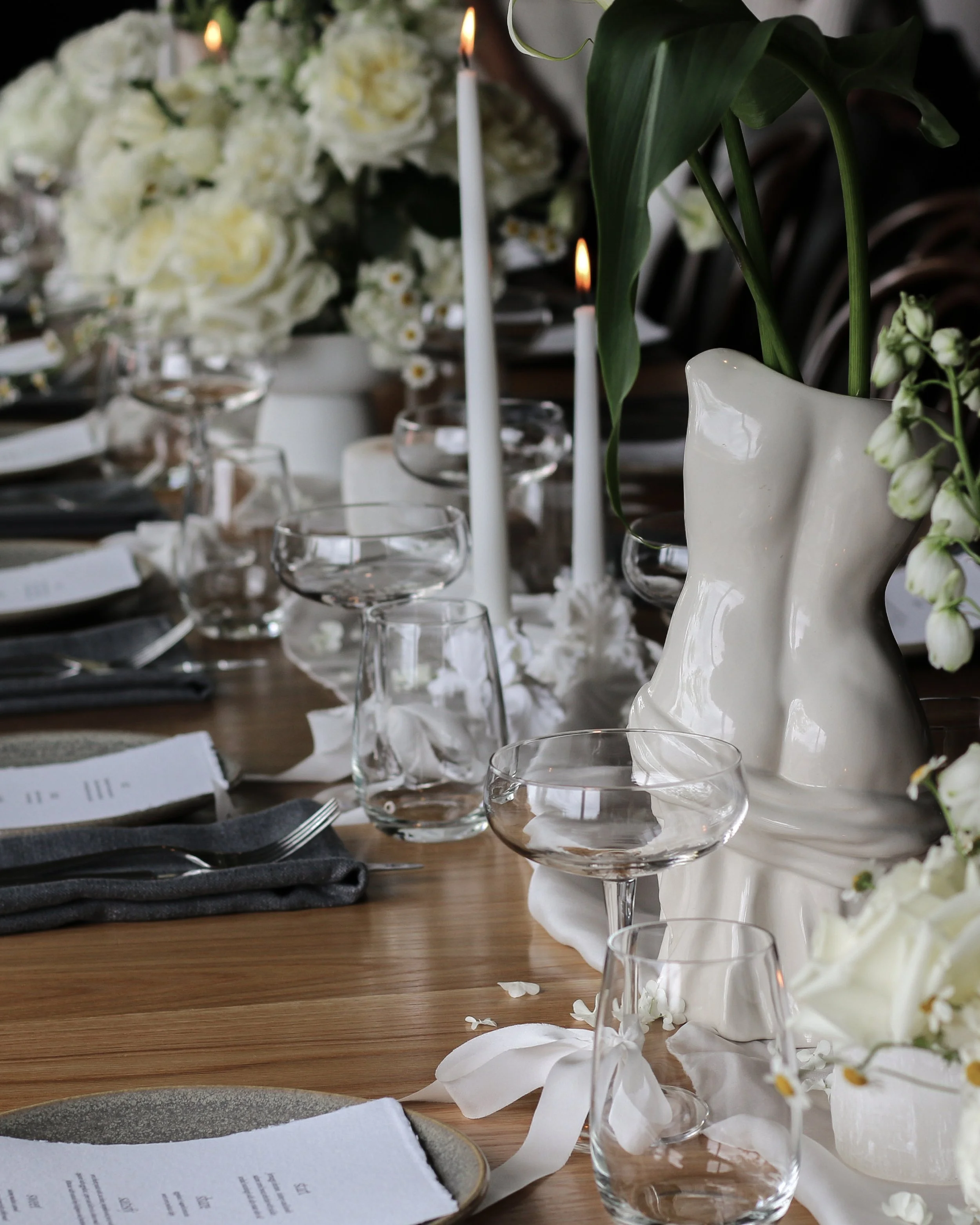 Elegant table setting with white flowers, candles, and a sculptural vase on a wooden table.