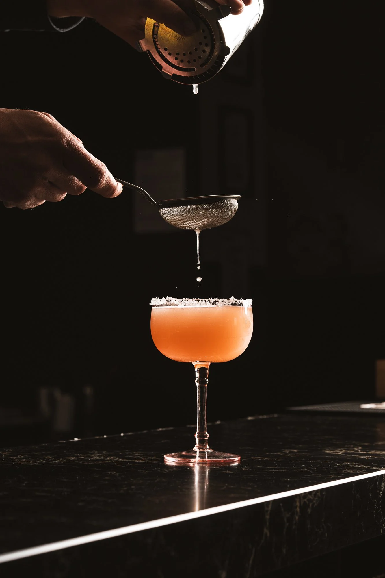 A bartender is straining a delicious pink cocktail into a coupe glass, with a salted rim at Assaggini Restaurant & Wine Bar.