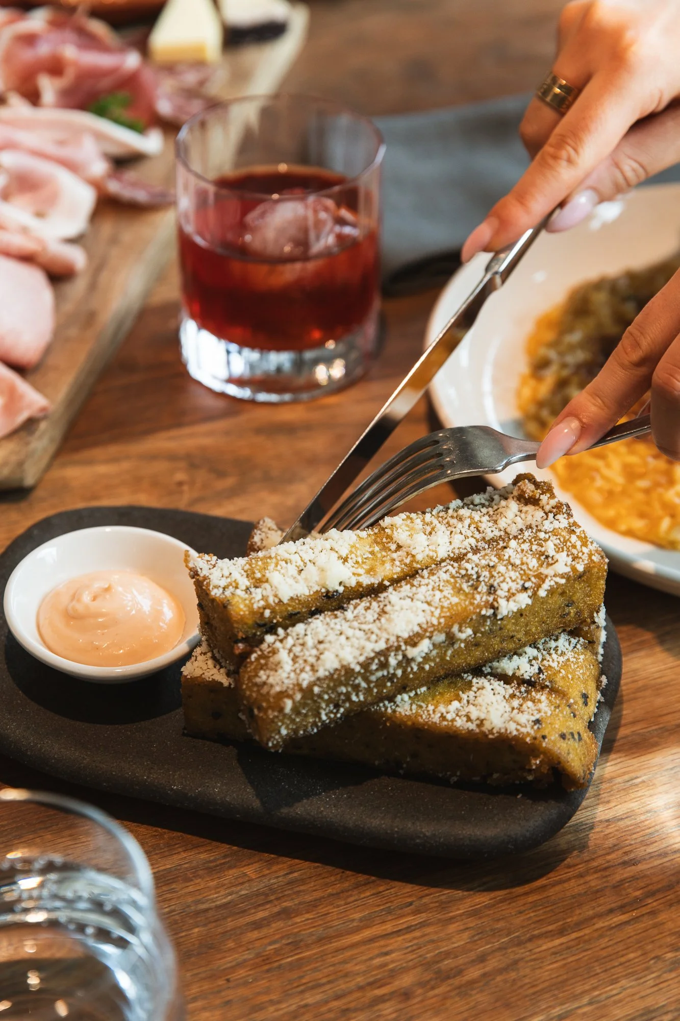 Stack of fried Polenta Chips sprinkled with Parmesan cheese served with dipping sauce on a black plate, with a glass of red drink and a plate of food in the background.