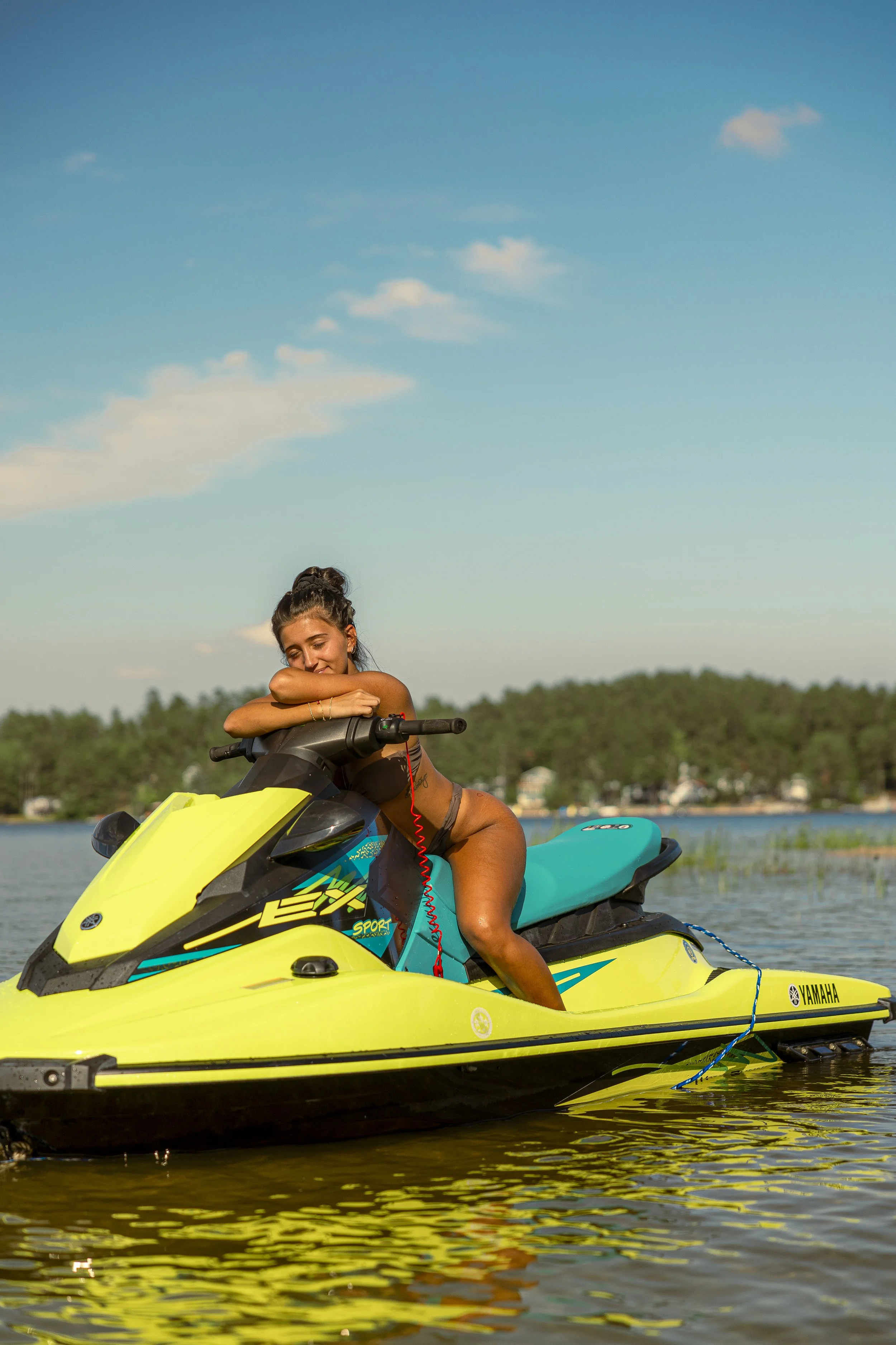 A woman hugging a yellow and black Yamaha jet ski on a lake, with trees and a blue sky in the background.