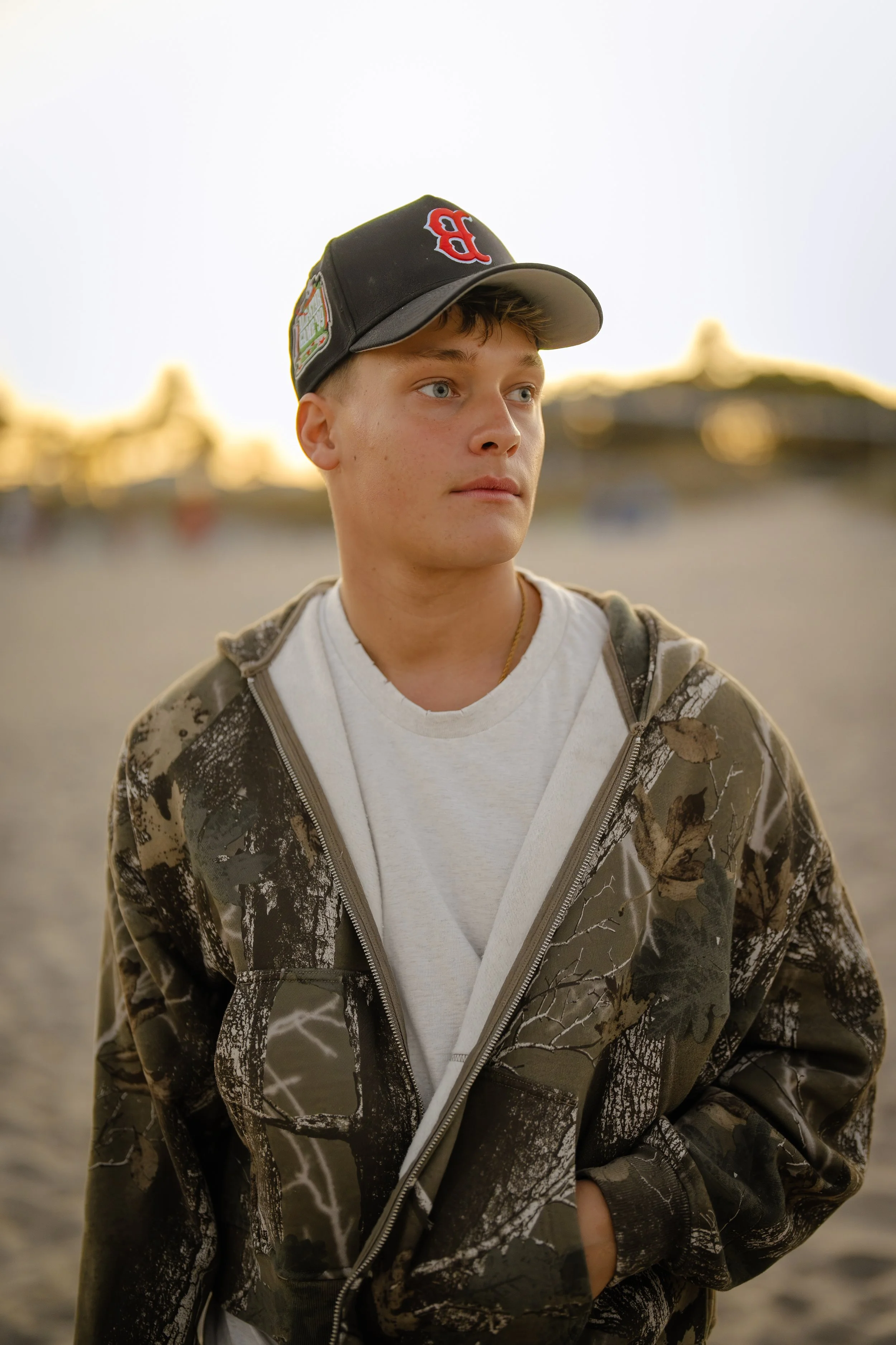 Young man wearing a camo jacket and a black baseball cap standing on a beach during sunset