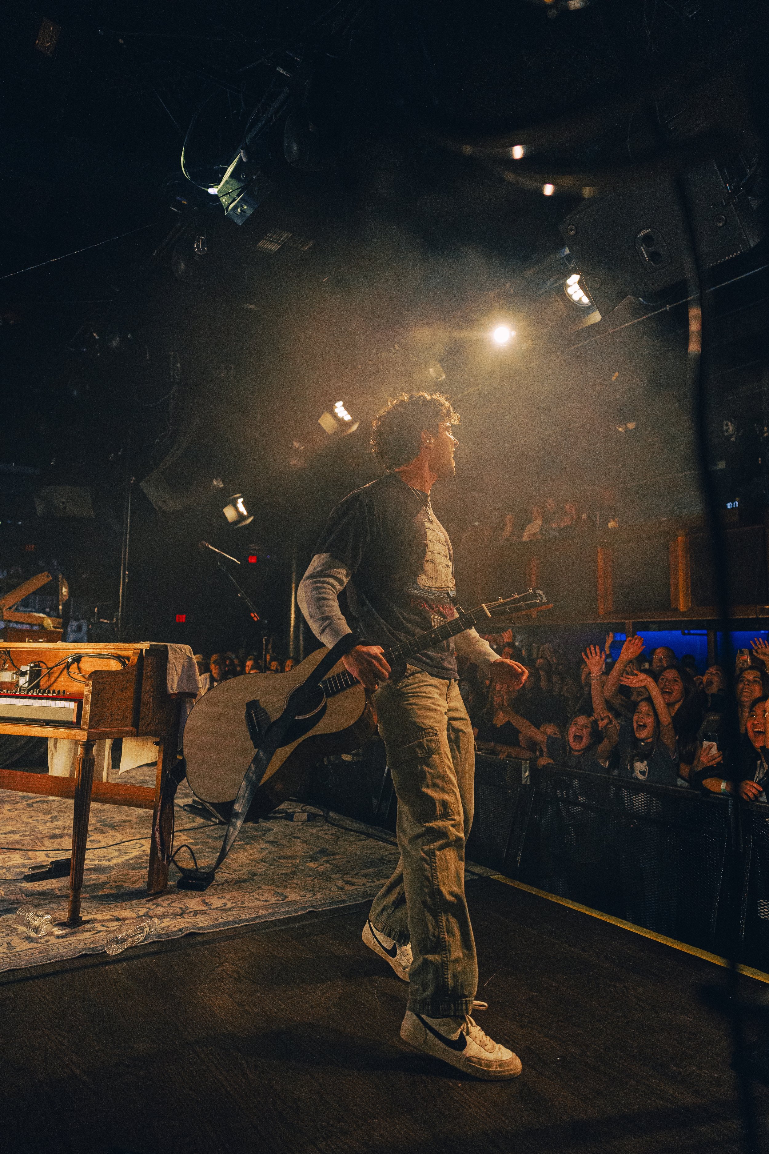 A young male musician holding an acoustic guitar stands on stage during a concert, facing a cheering audience with some fans raising their hands.