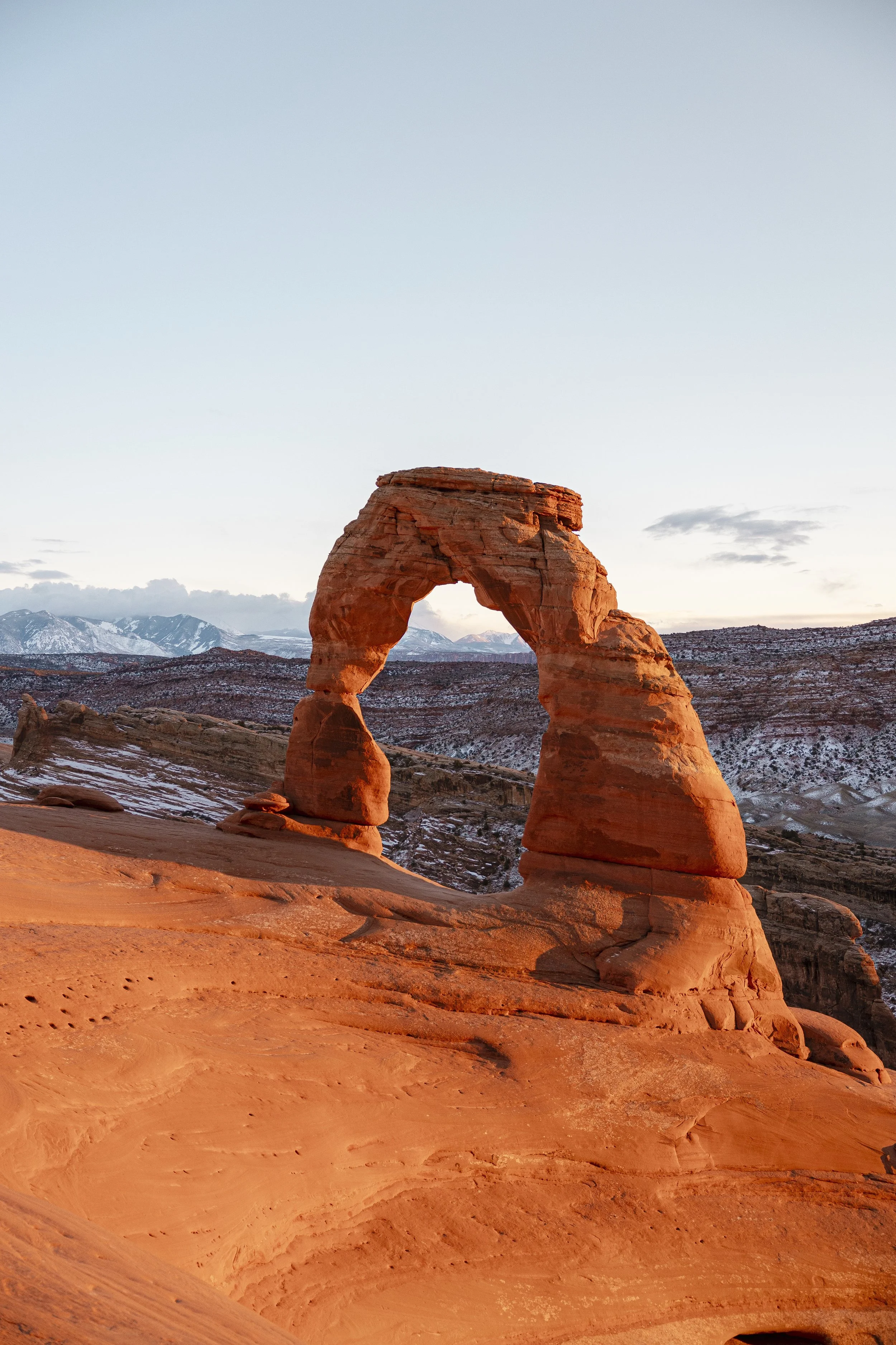 Sunset over desert landscape featuring the iconic Delicate Arch formation in Arches National Park, Utah, with snow-capped mountains in the background.