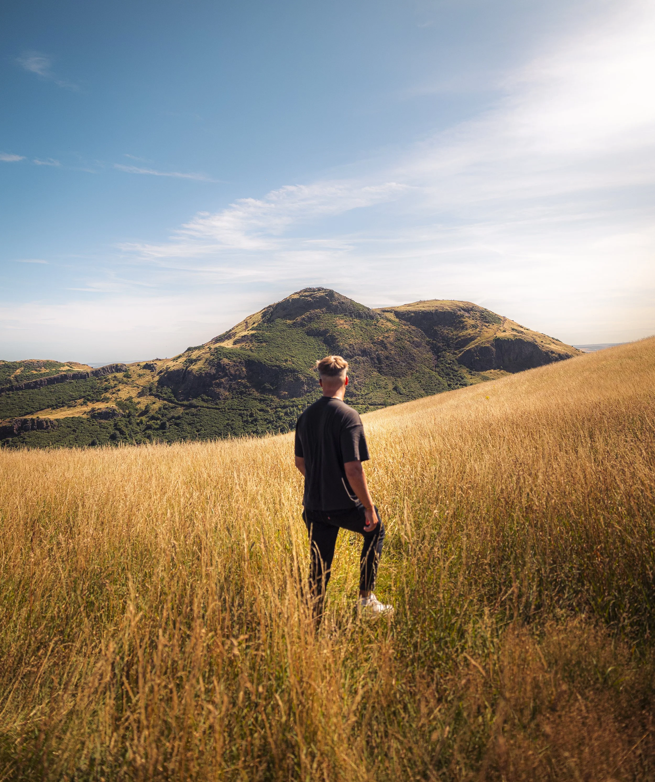 Man standing in a grassy field with green hills and mountains in the background under a partly cloudy sky.
