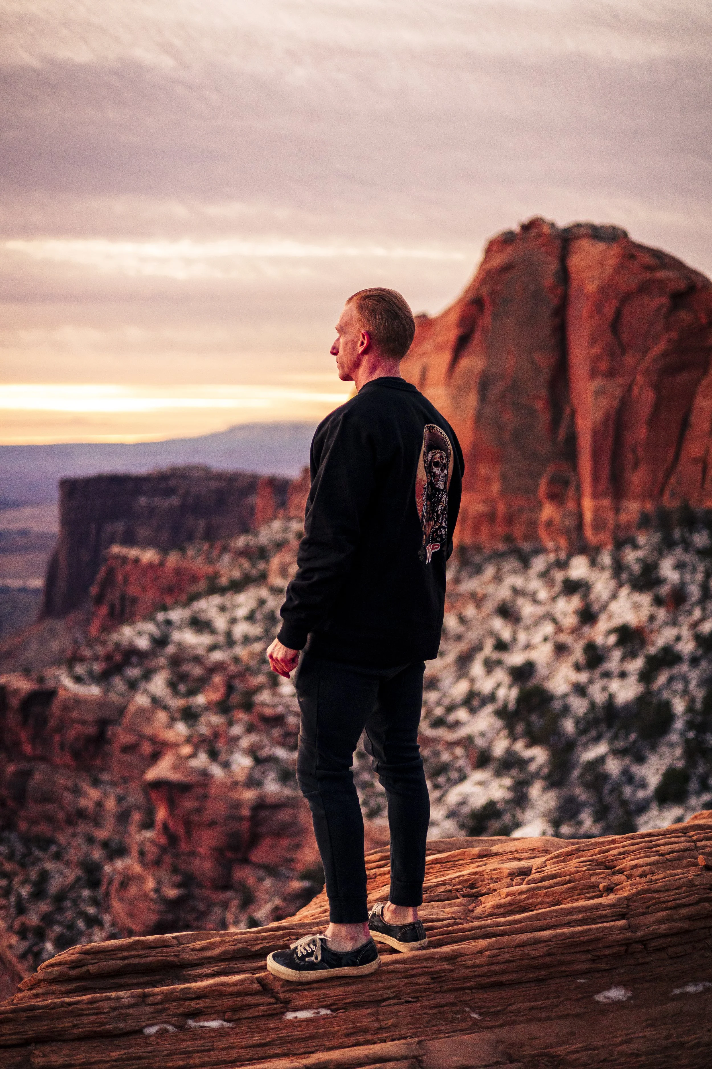 A man standing on a rock ledge overlooking a desert canyon at sunset, wearing a black jacket, black pants, and sneakers.