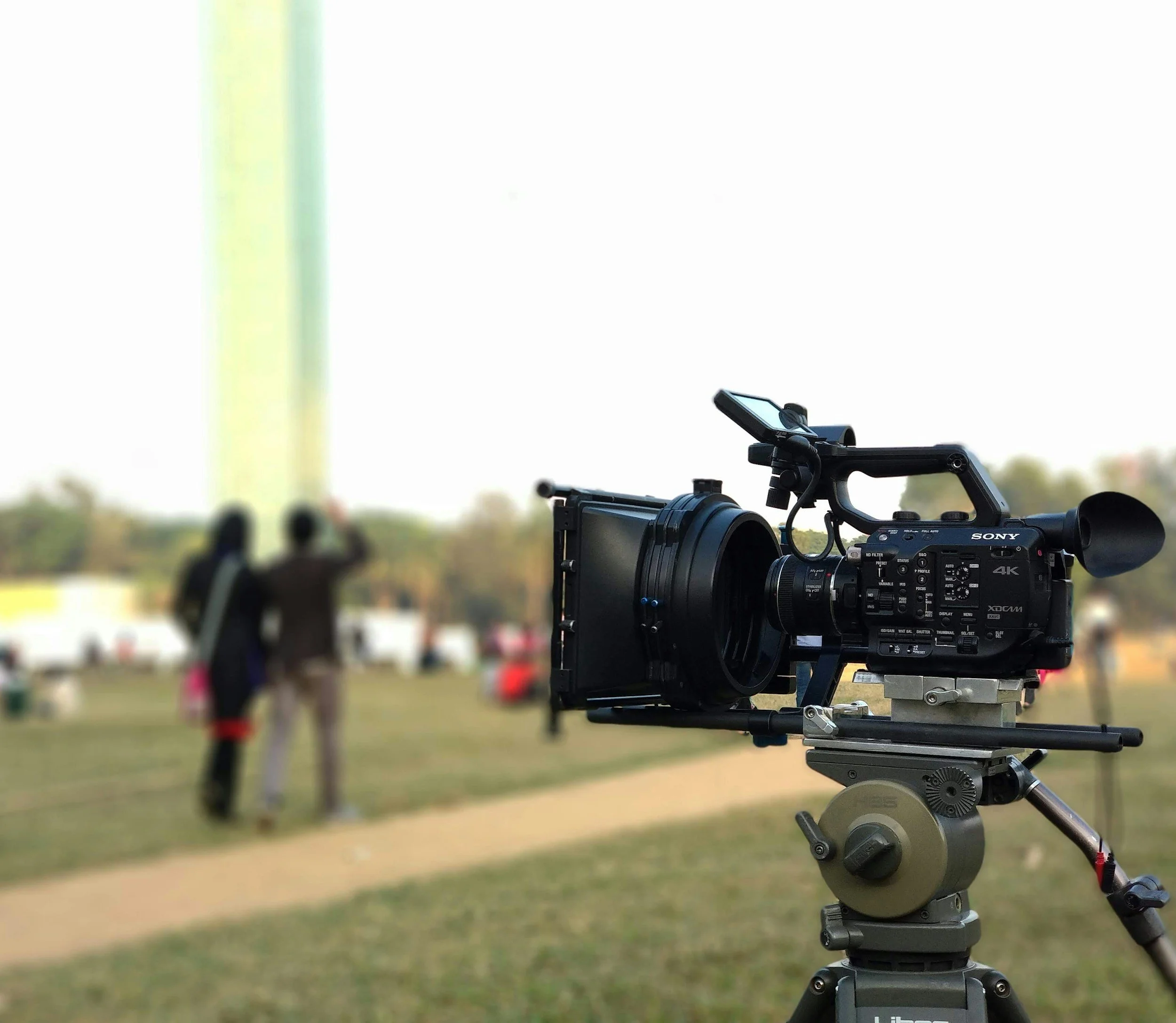 Professional video camera on tripod filming outdoors with blurred background of people and trees.