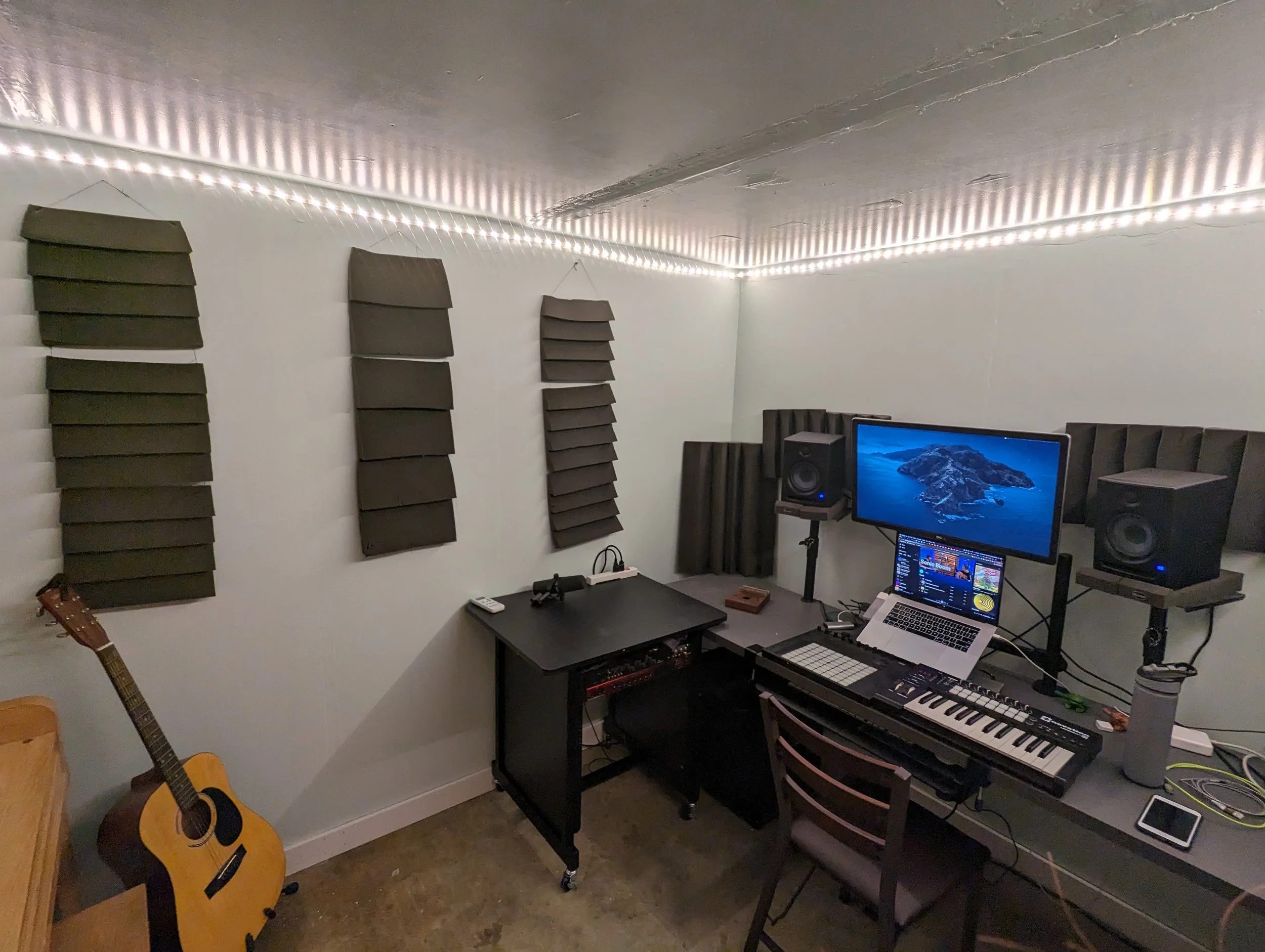 Music studio with acoustic foam panels on the walls, a guitar, a computer monitor, speakers, a keyboard, and a microphone setup.