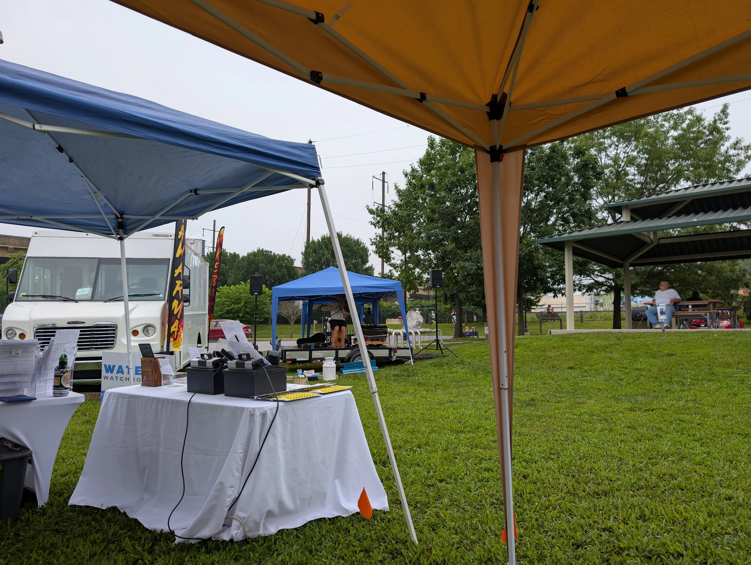 Outdoor event setup with tents, tables, and a food truck on a grassy field. People are seen sitting and walking around under trees and shelter, with a man sitting on a bench in the background.