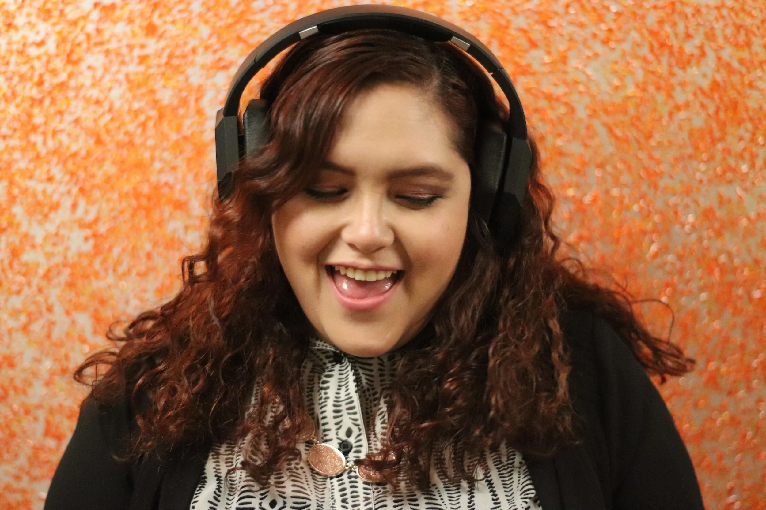 Woman with curly hair wearing headphones. She is facing slightly down with her mouth in an open smile