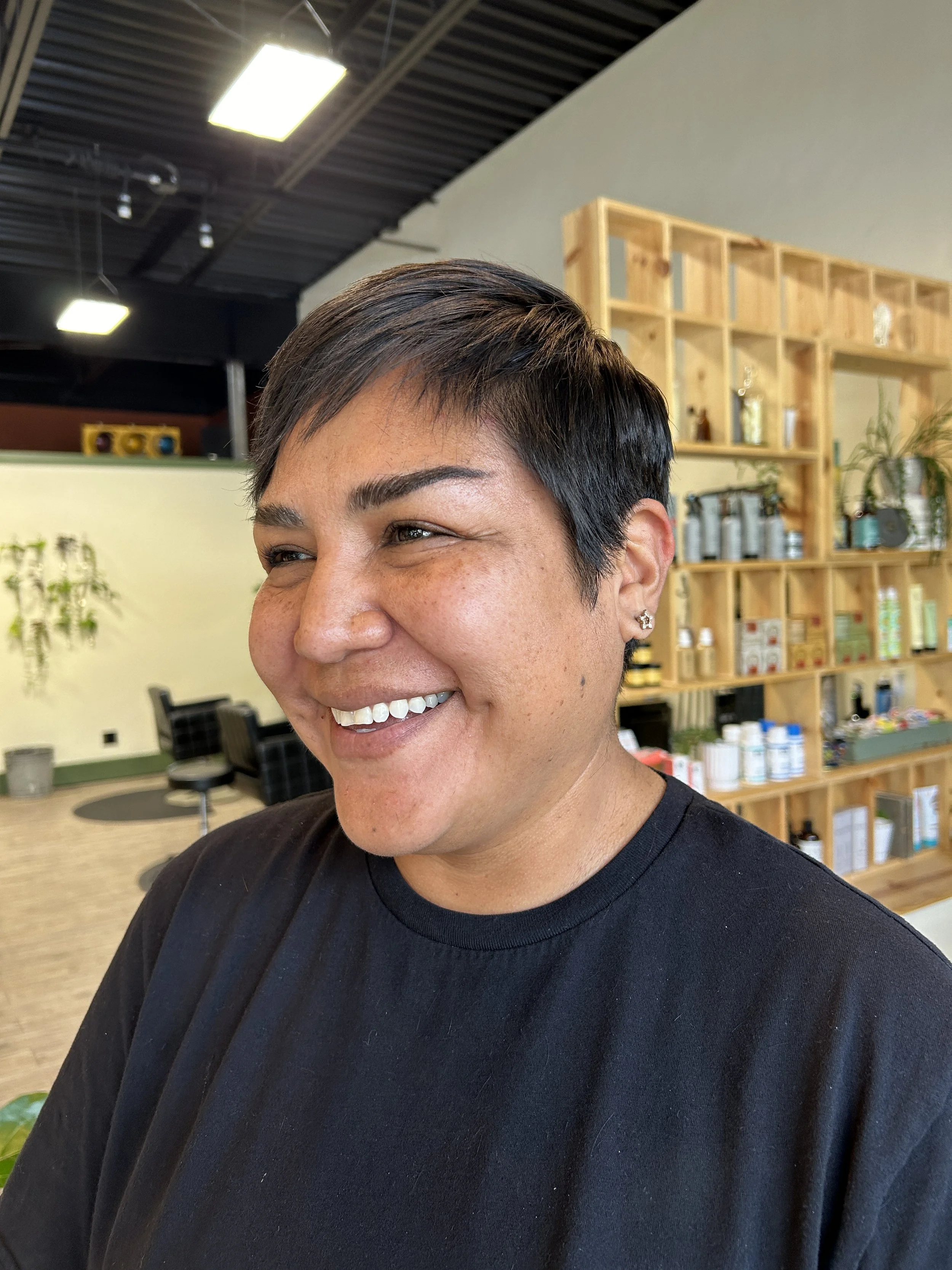 A smiling woman with short dark hair and earrings in a salon with wooden shelving and products in the background.