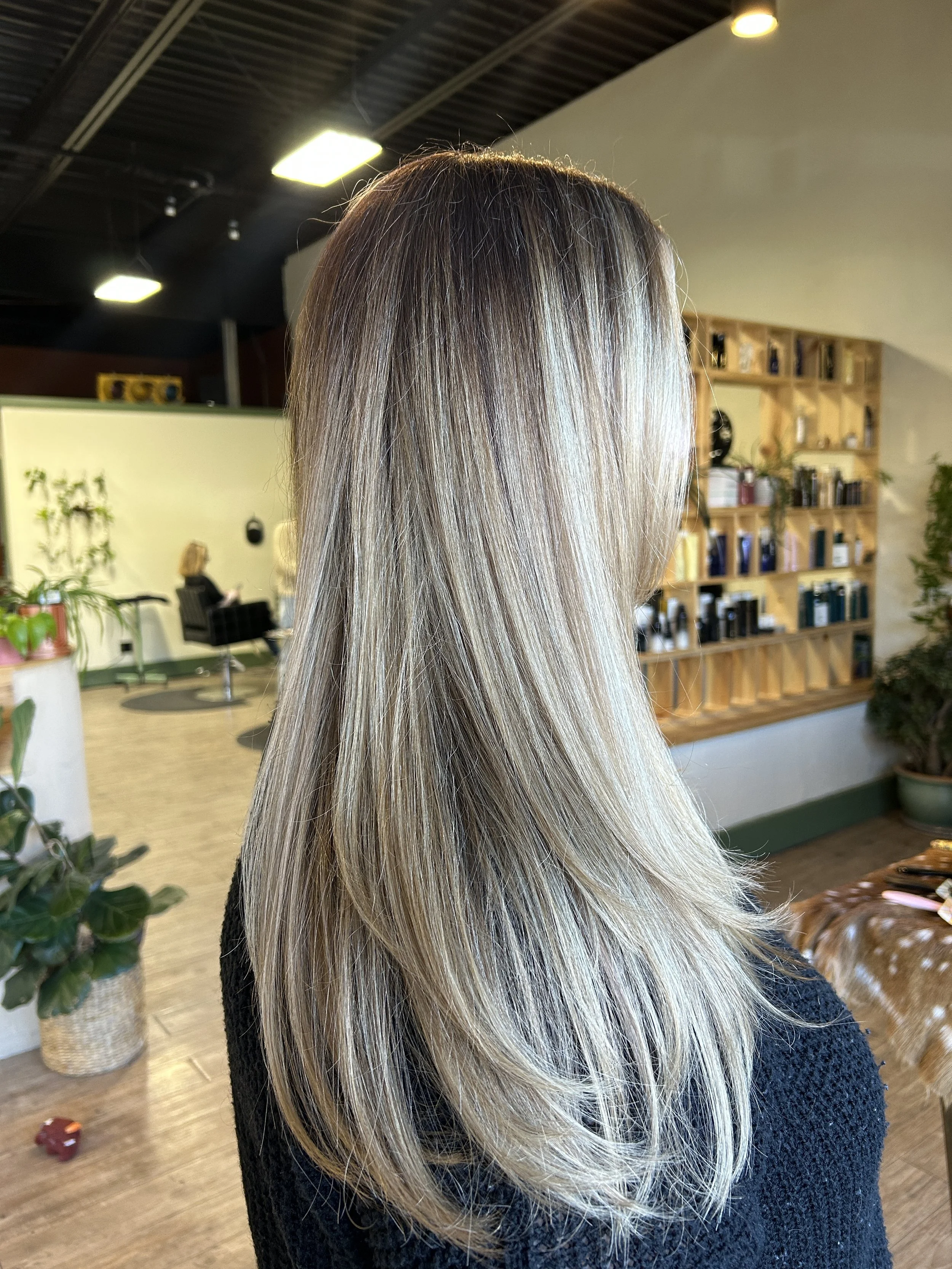 Side view of a woman with long, straight blonde hair, sitting inside a hair salon with wooden shelves and potted plants in the background.