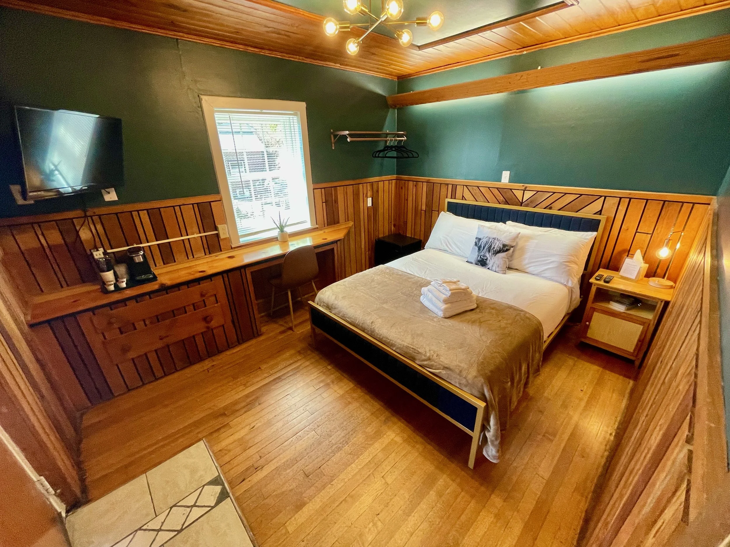 A cozy bedroom with wood paneled walls and a large bed with white bedding. There are towels folded on the bed, a nightstand with a lamp and phone, a small window with a plant on the desk below, a wall-mounted TV, and a ceiling light fixture with multiple bulbs.