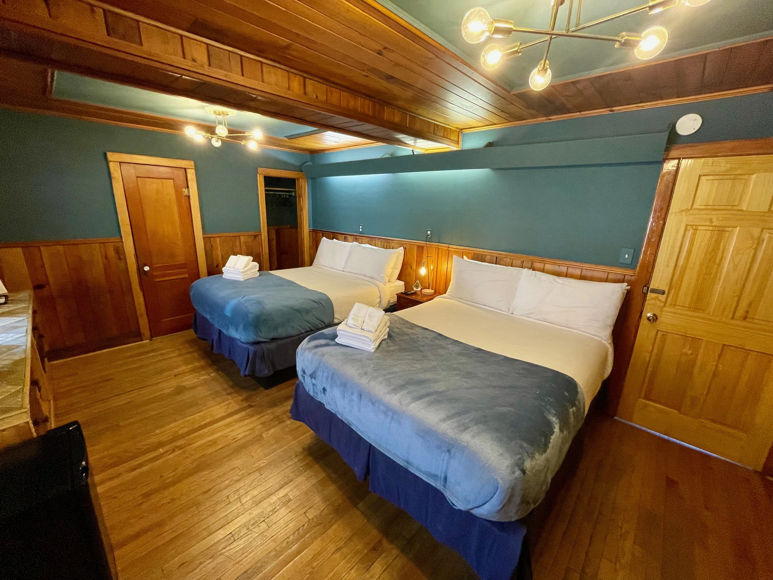 Two queen beds in a cozy hotel room with wooden wall paneling and blue walls, bedside table with lamp, hardwood floors, wooden doors, and modern ceiling lights.