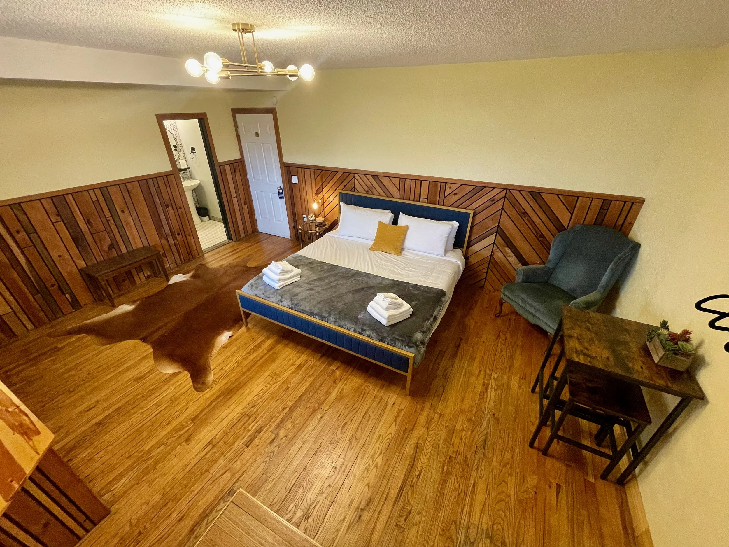 A cozy bedroom with wooden flooring, a blue bed, a pair of folded white towels on the bed, a dark green armchair, a small wooden table with a flower arrangement, and an oval cowhide rug. There is a door leading to a bathroom and a modern ceiling light fixture.