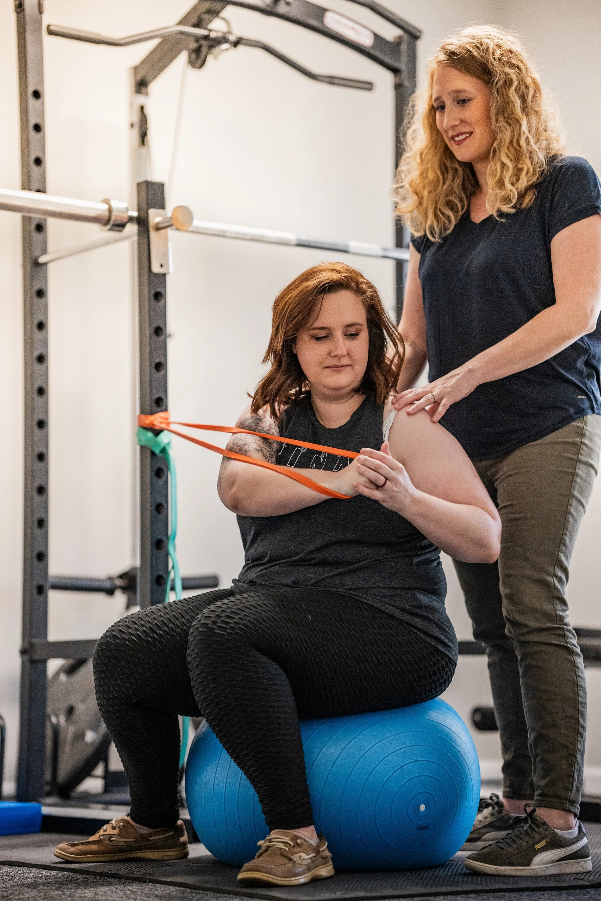 Dr. Noelle, a woman with curly hair supervises as a patient, a woman wearing a tank top and with visible tattoos sits on an exercise ball and pulls on a band. In the back ground you can see a squat rack.