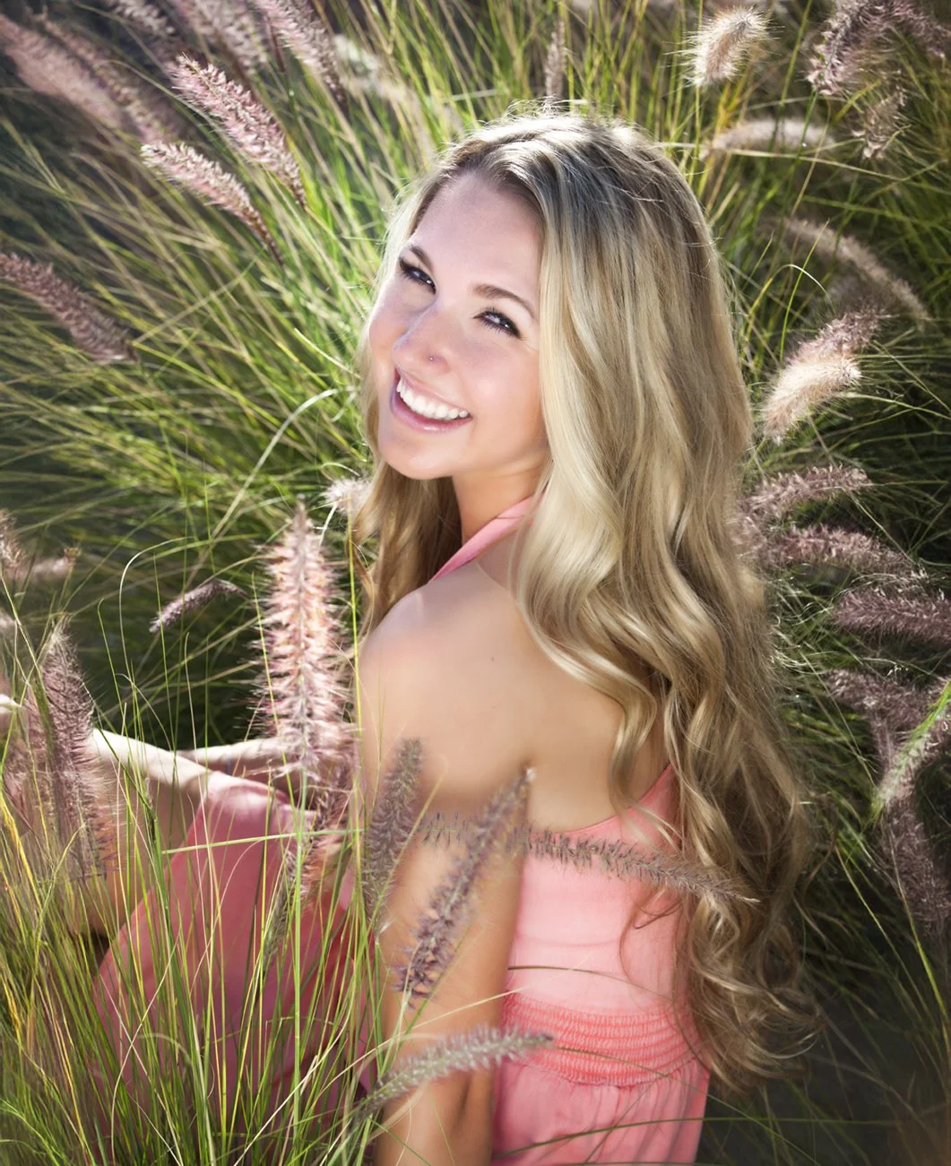 A smiling senior woman with long wavy blonde hair in a pink dress among tall grass with feathery purple flowers. Senior photos in Tempe Arizona.