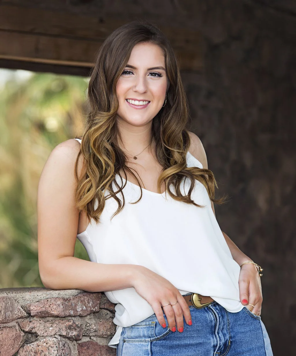 A young high school senior woman with long brown hair and light skin, wearing a white sleeveless top and blue jeans, smiling outdoors with a blurred natural background.