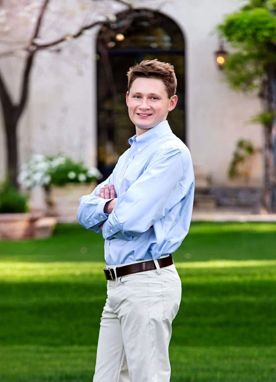 A senior male with short brown hair and fair skin standing outdoors on a well-maintained lawn, with his arms crossed, smiling and looking at the camera, in front of a building with a large arched window and greenery. High School Senior.