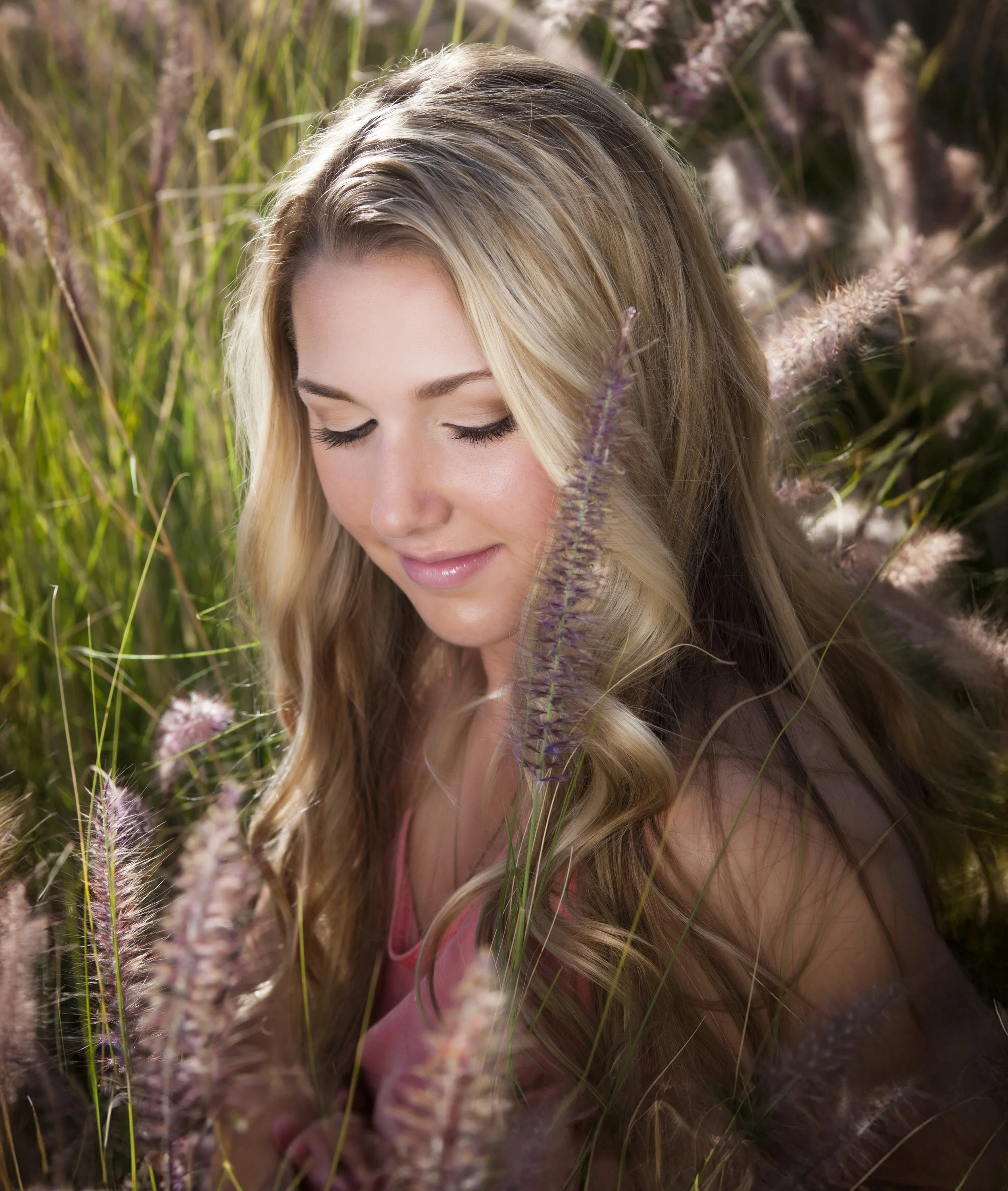 A young woman with long blonde hair and closed eyes sitting among tall grasses and flowers in a natural outdoor setting. Senior graduation photos.
